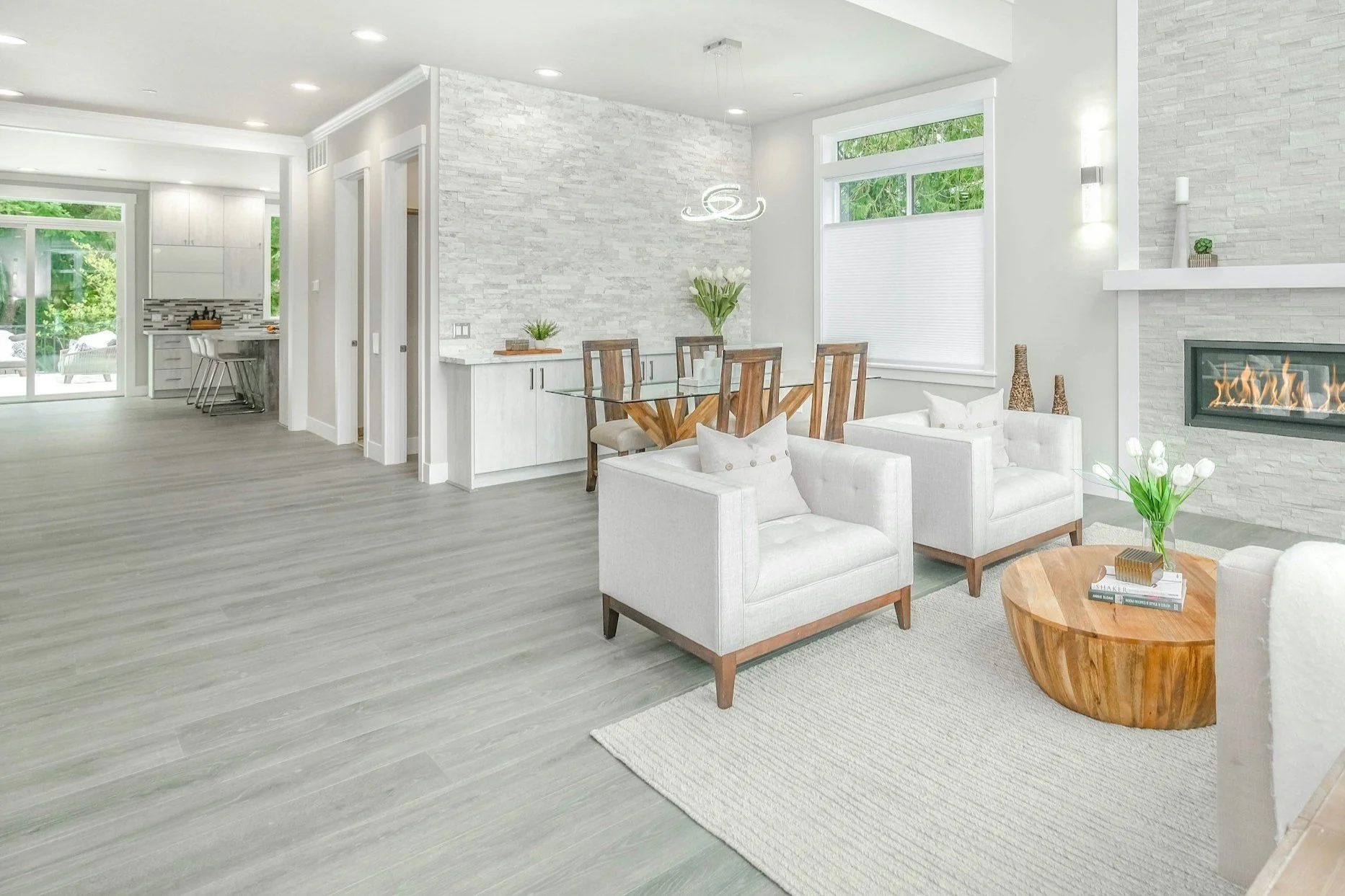 Sala de estar moderna con chimenea, muebles de color claro, mesa de centro de madera, arreglos de flores, comedor al fondo y piso de madera clara.