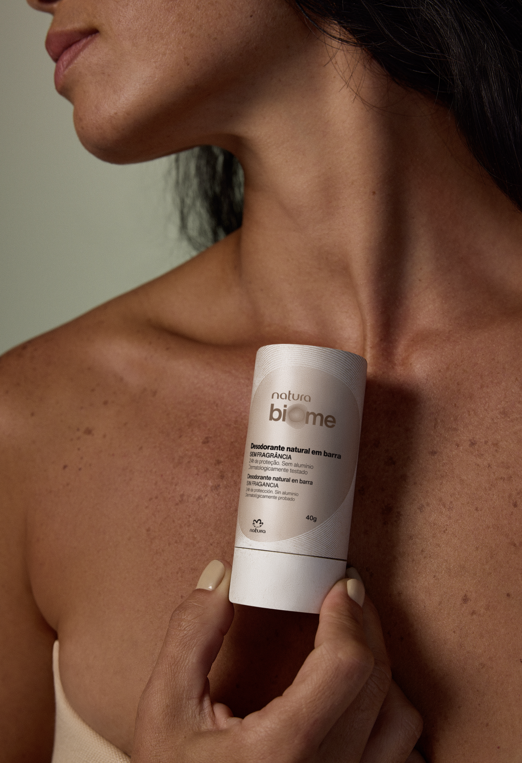 Close-up of a woman with tanned skin holding a tube of natural deodorant stick at neck level, showing the product with close-up details of the packaging.