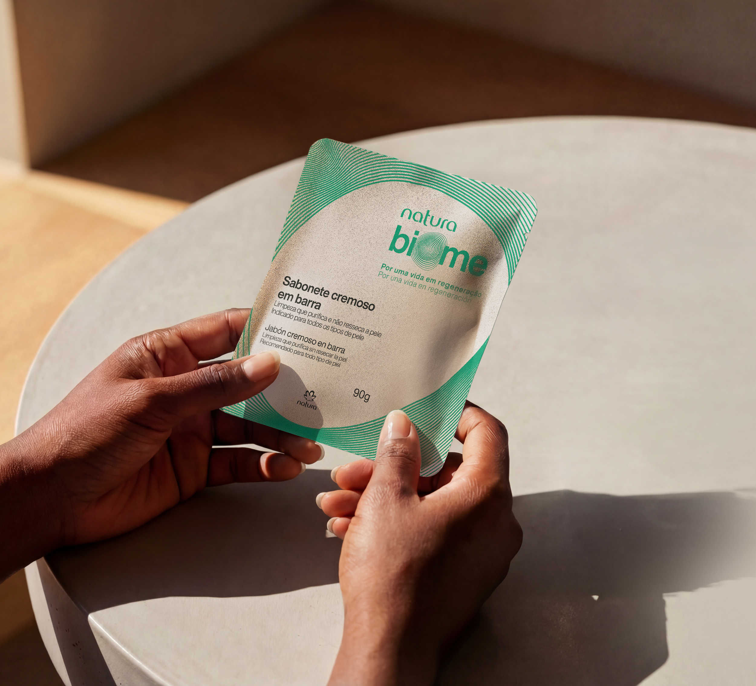 Hands holding a bar of soap in Natura Biome packaging, on a white table.