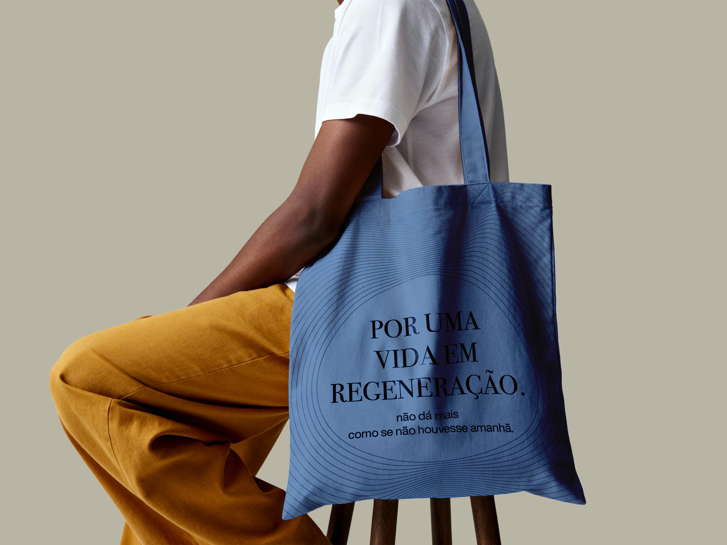 A person sitting in a wooden chair, wearing mustard-colored pants and a white T-shirt, holding a blue bag with a message written on it.