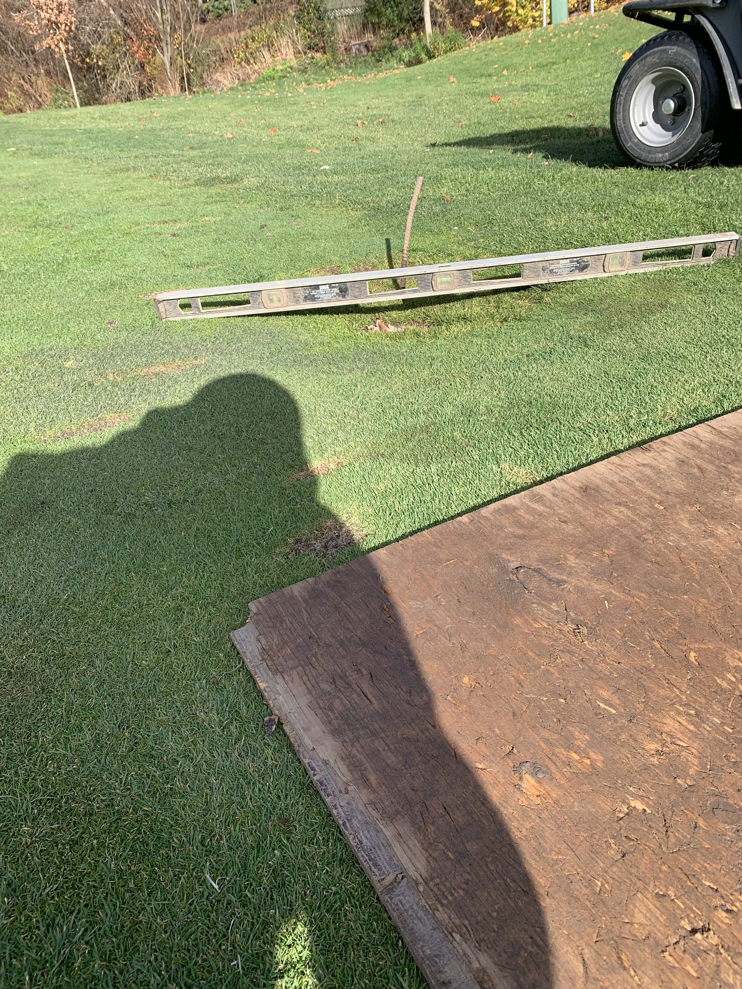 A level tool, a piece of wood, and a part of a black vehicle tire on a grassy area, with a shadow of a person taking the photo. picture shows dip in ground as ground of sprinkler has sunk