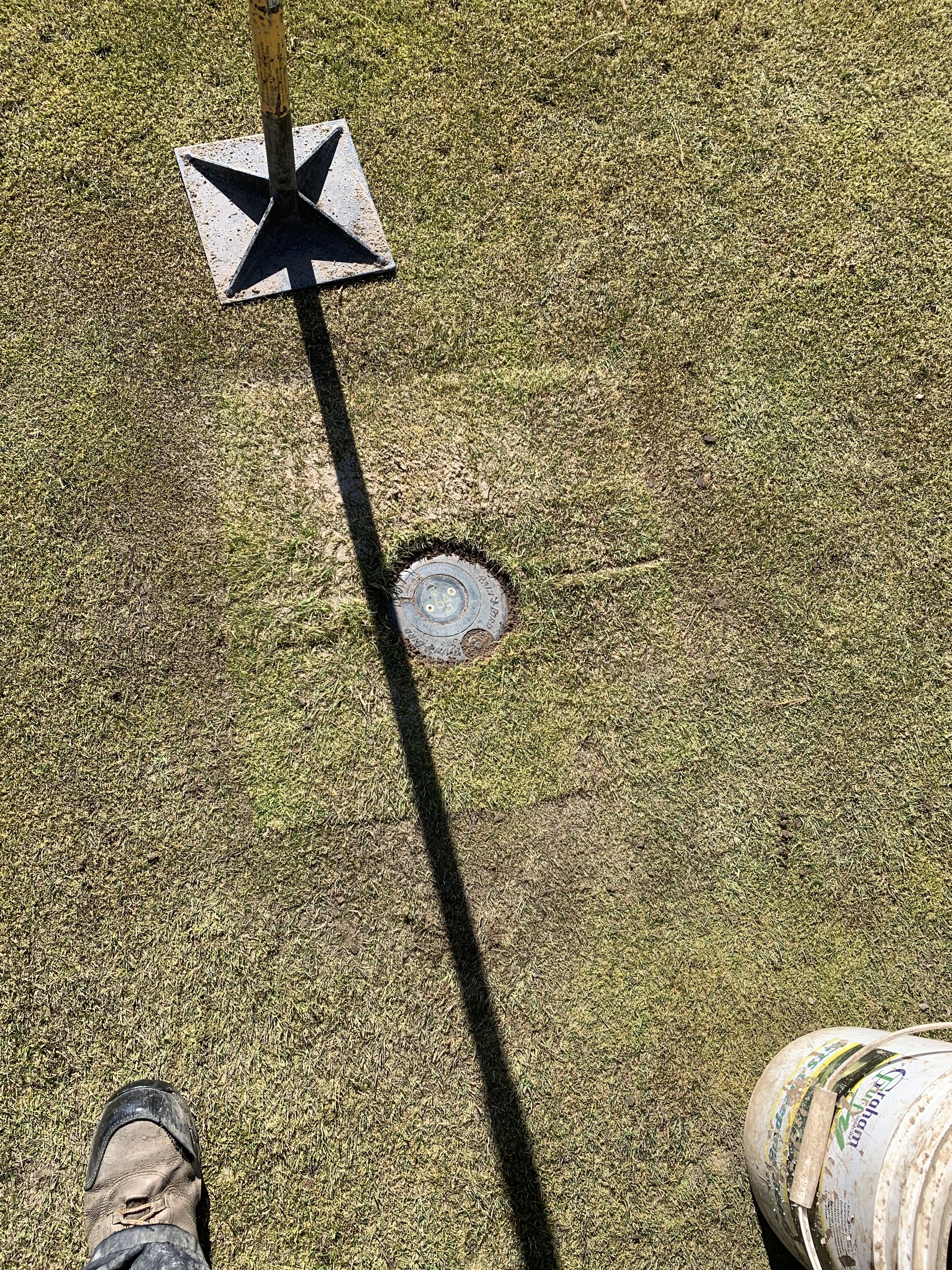A golf course with a golf flag and hole. The flag is casting a shadow. Part of a person's shoe and a bucket of golf balls are visible in the bottom corners.