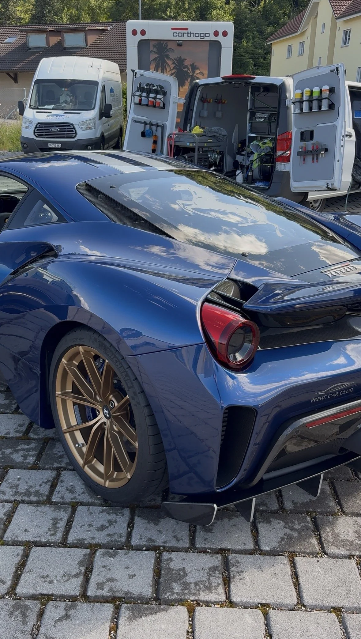 Ein blauer Sportwagen, vermutlich Ferrari, steht auf einem gepflasterten Platz. Im Hintergrund sind zwei Servicefahrzeuge aufgestellt, eines mit geöffneten Türen, die Werkzeug und Verbrauchsmaterialien zeigen.