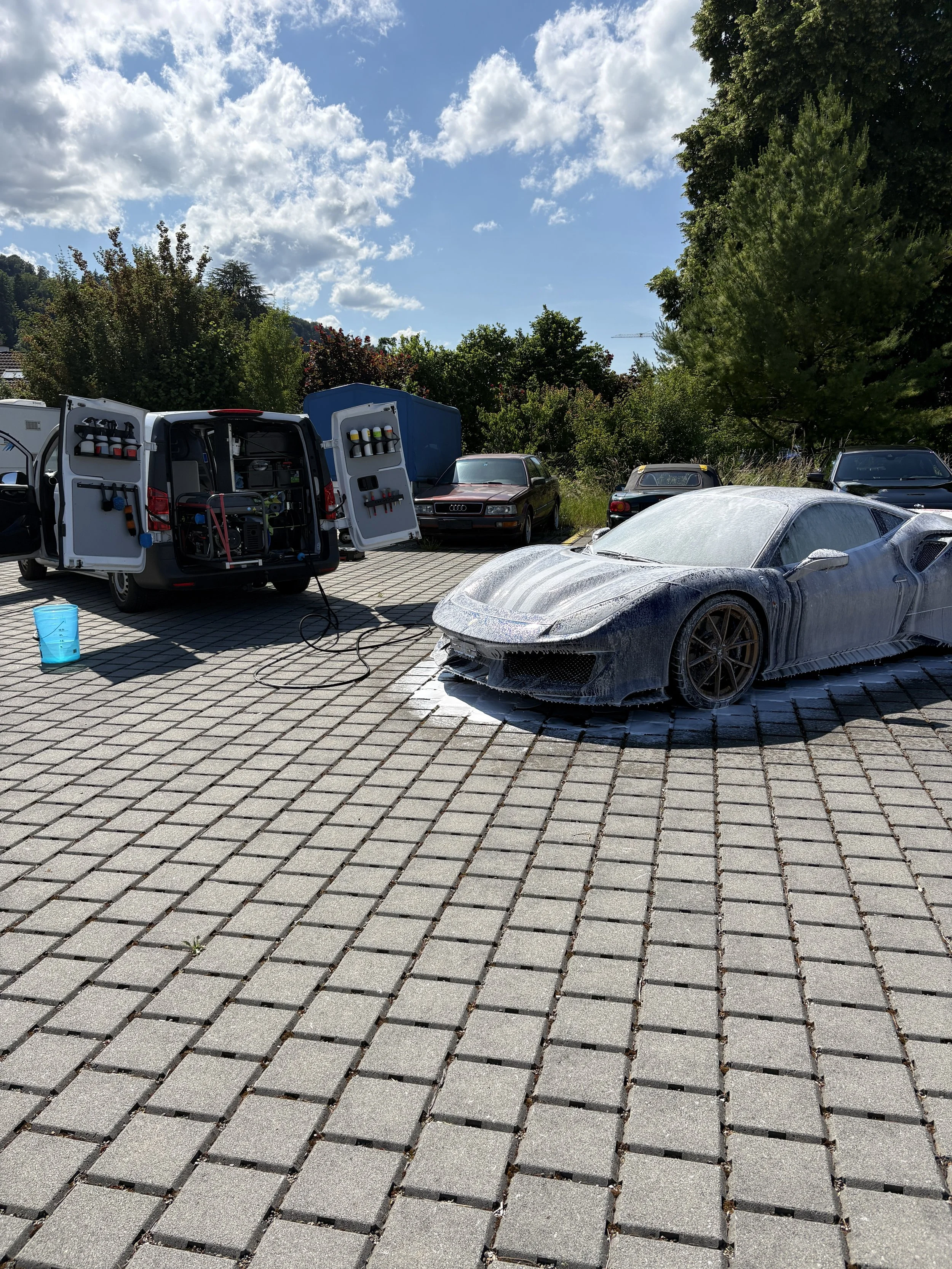 Ein angetriebener Sportwagen wird mit Seifenwasser gereinigt, im Hintergrund stehen Autos auf einem gepflasterten Parkplatz, umgeben von Bäumen und einem blauen Himmel mit Wolken.
