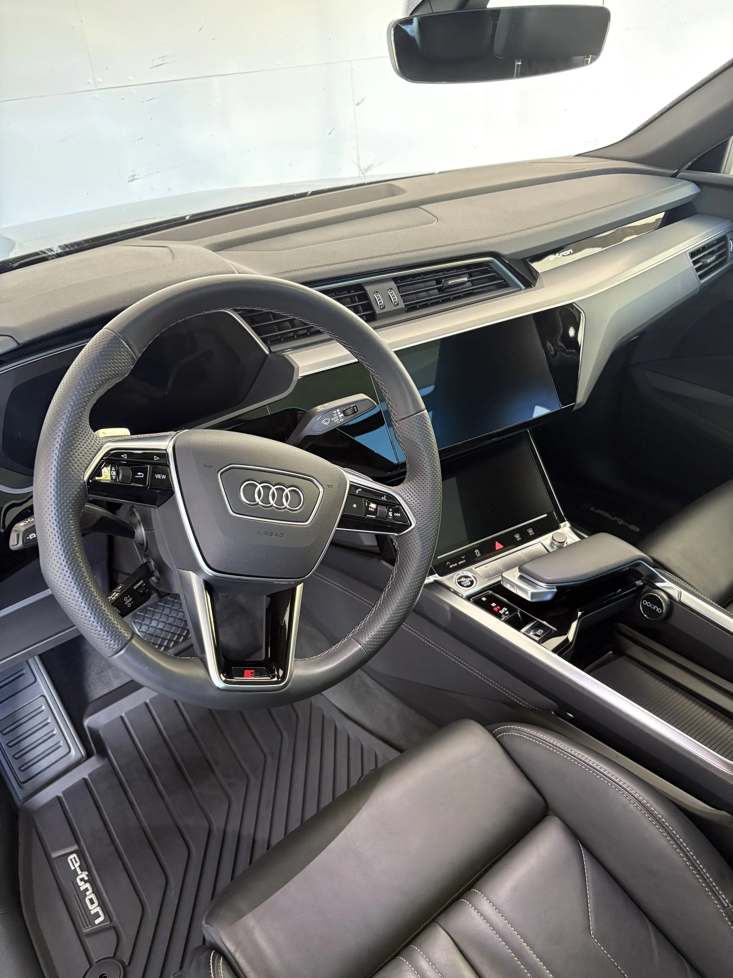Innenraum eines Audi e-tron Fahrzeugs, sicht auf das Lenkrad, Mittelkonsole mit Touchscreens und Ablagefläche, hochwertige schwarze Ledersitze, Gummimatten auf dem Boden.
