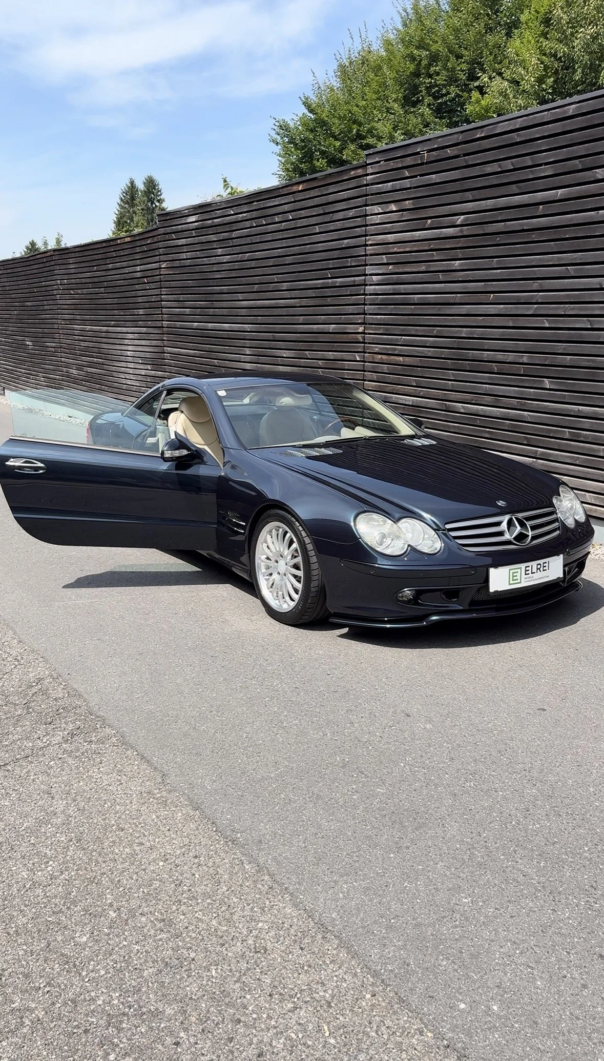 Ein schwarzer Mercedes-Benz Sportwagen mit geöffnetem Fahrerhausschnitt, parkend vor einer hölzernen Mauer, während die Umgebung grün und sonnig ist.
