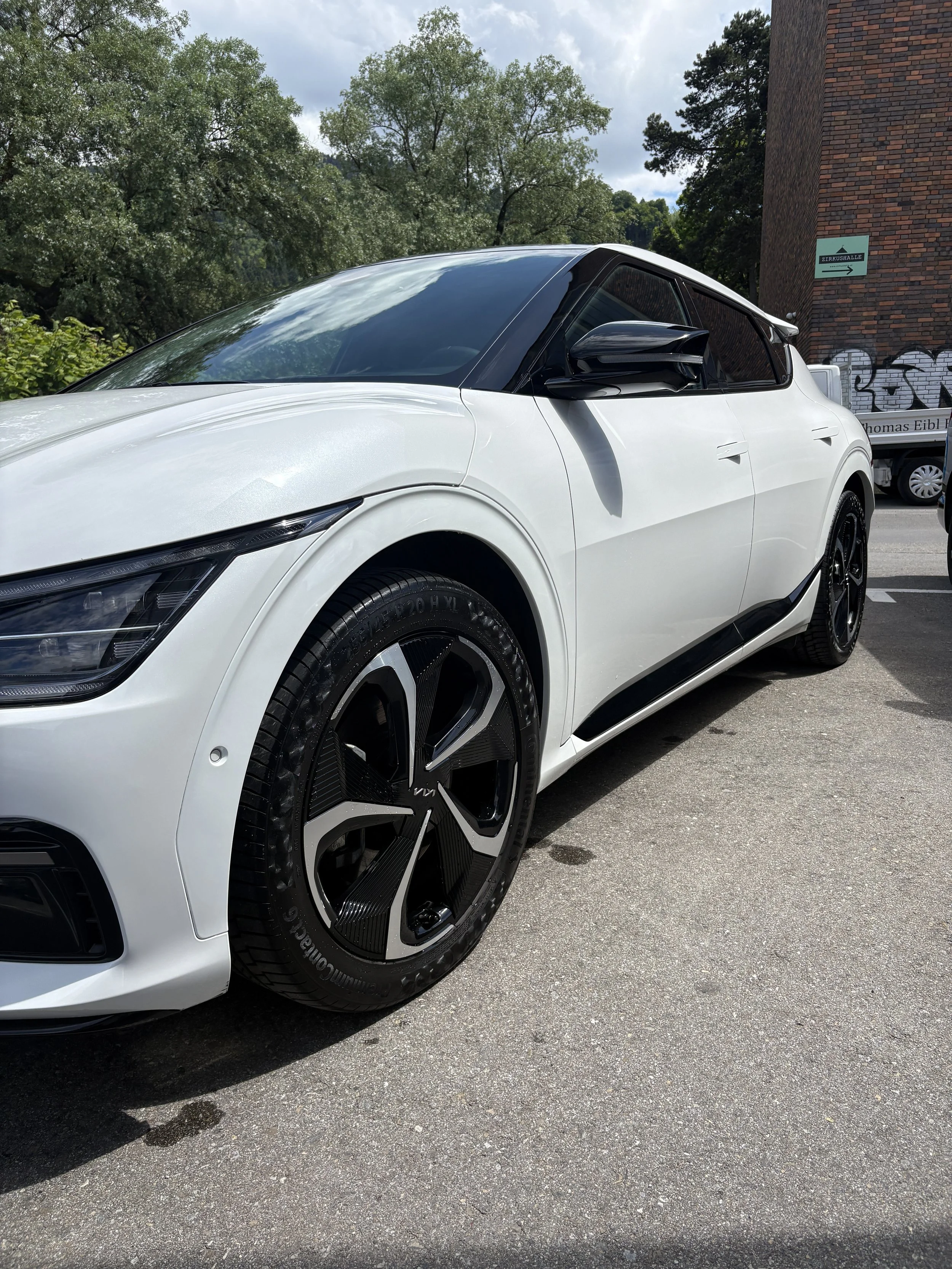 Weiße Sportlimousine mit schwarzen Akzenten und großen Radialreifen, parkend auf einer asphaltierter Straße, im Hintergrund Bäume und ein Gebäude mit einem Graffiti