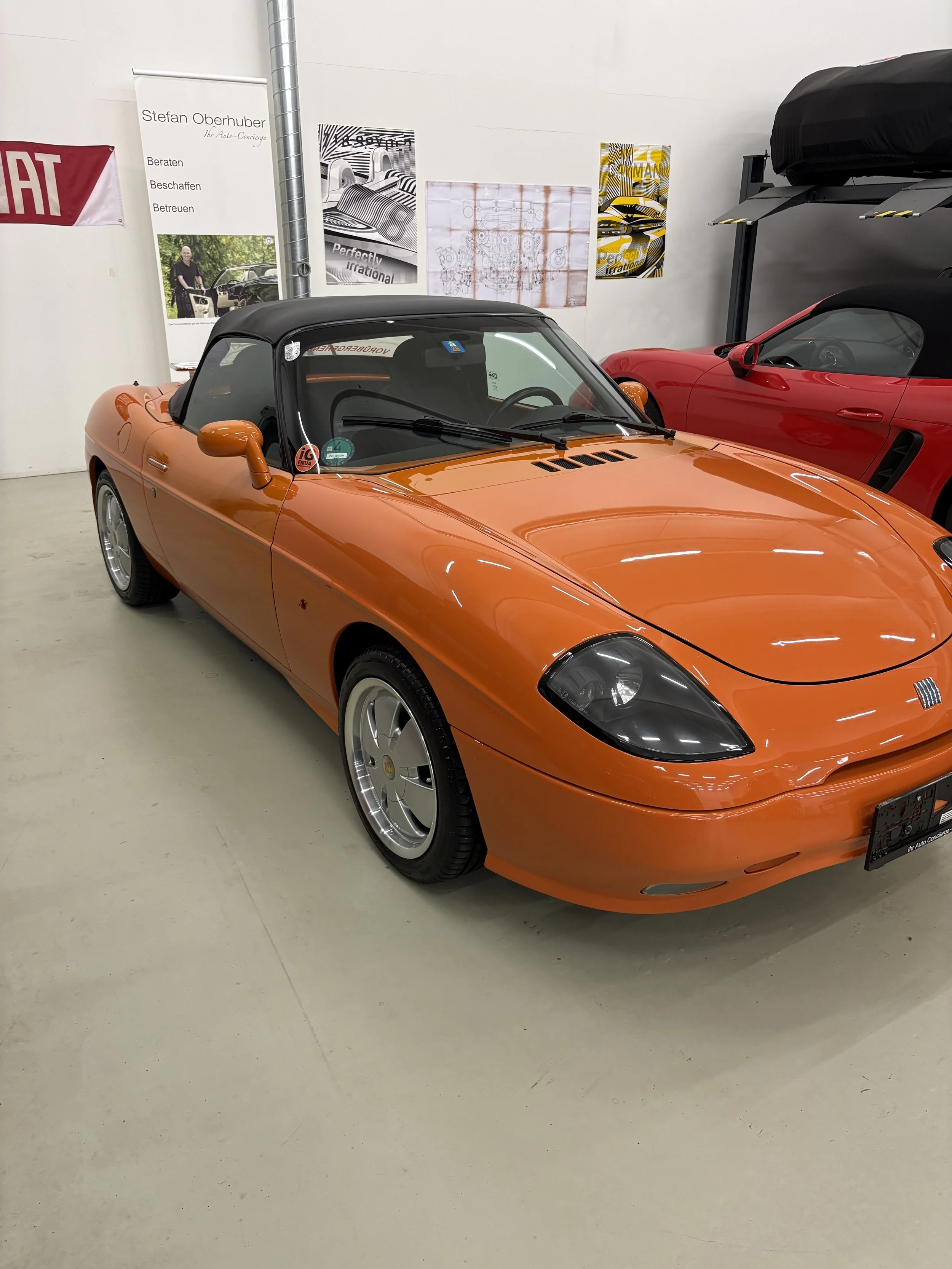 Ein orangefarbener Oldtimer mit Softtop auf einer hellen, glatten Bodenfläche in einer Ausstellungshalle. Im Hintergrund stehen Poster und ein anderes rotes Auto. An der Wand hängt ein Banner mit dem Schriftzug "Stefan Oberhuber".