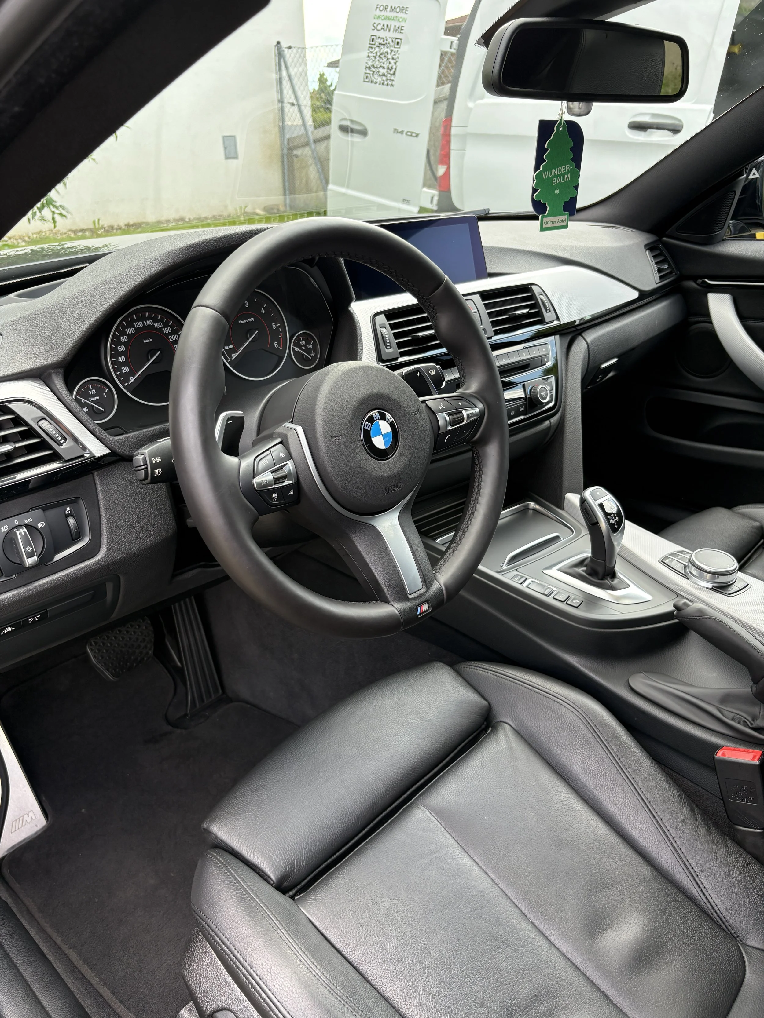 Innenraum eines BMW Fahrzeugs mit schwarzer Lederausstattung, einem Lenkrad mit BMW Logo, einem Schaltknüppel, einem Displaybildschirm und einer Fußmatte mit BMW Logo. Im Hintergrund sind die Rücksitze sichtbar.