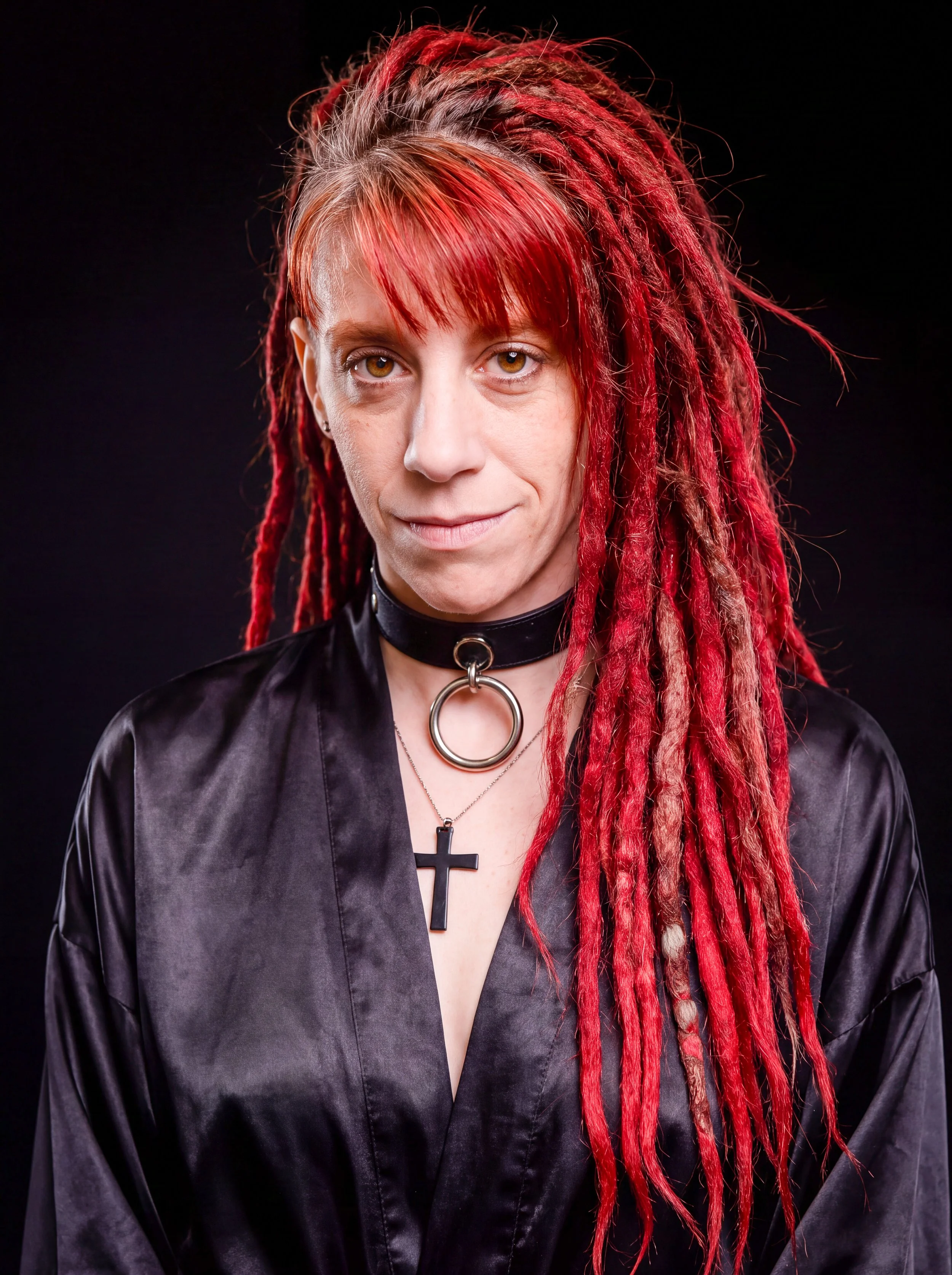 A woman with red dreadlocks, wearing a black leather jacket and cross necklace, posing against a black background.