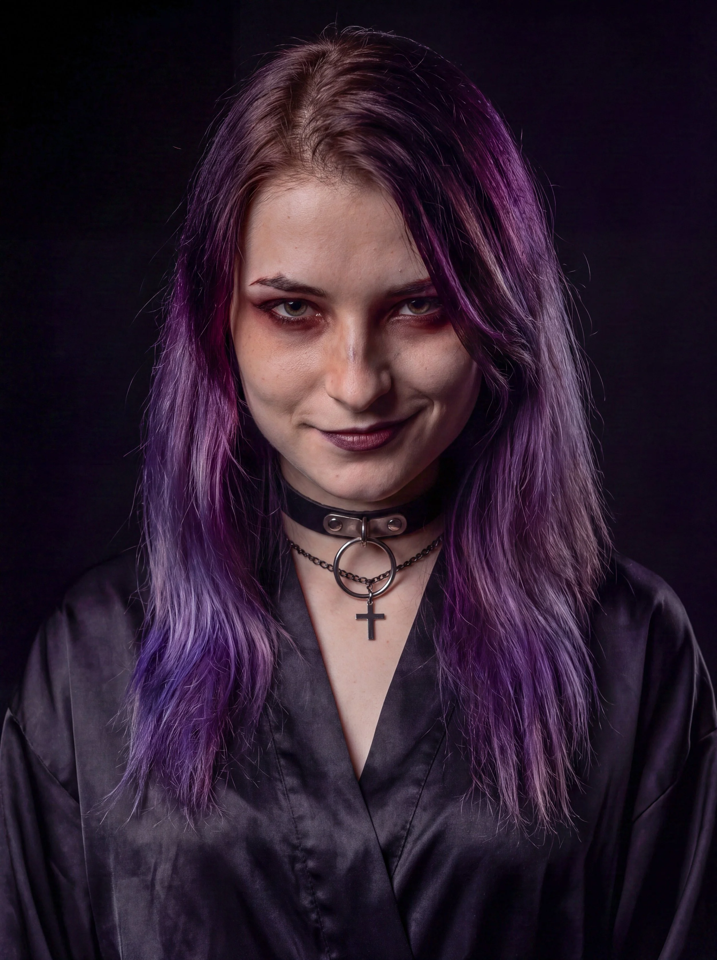 A woman with purple and pink hair wearing a choker with a cross pendant and a satin black shirt, smiling slightly against a dark background.