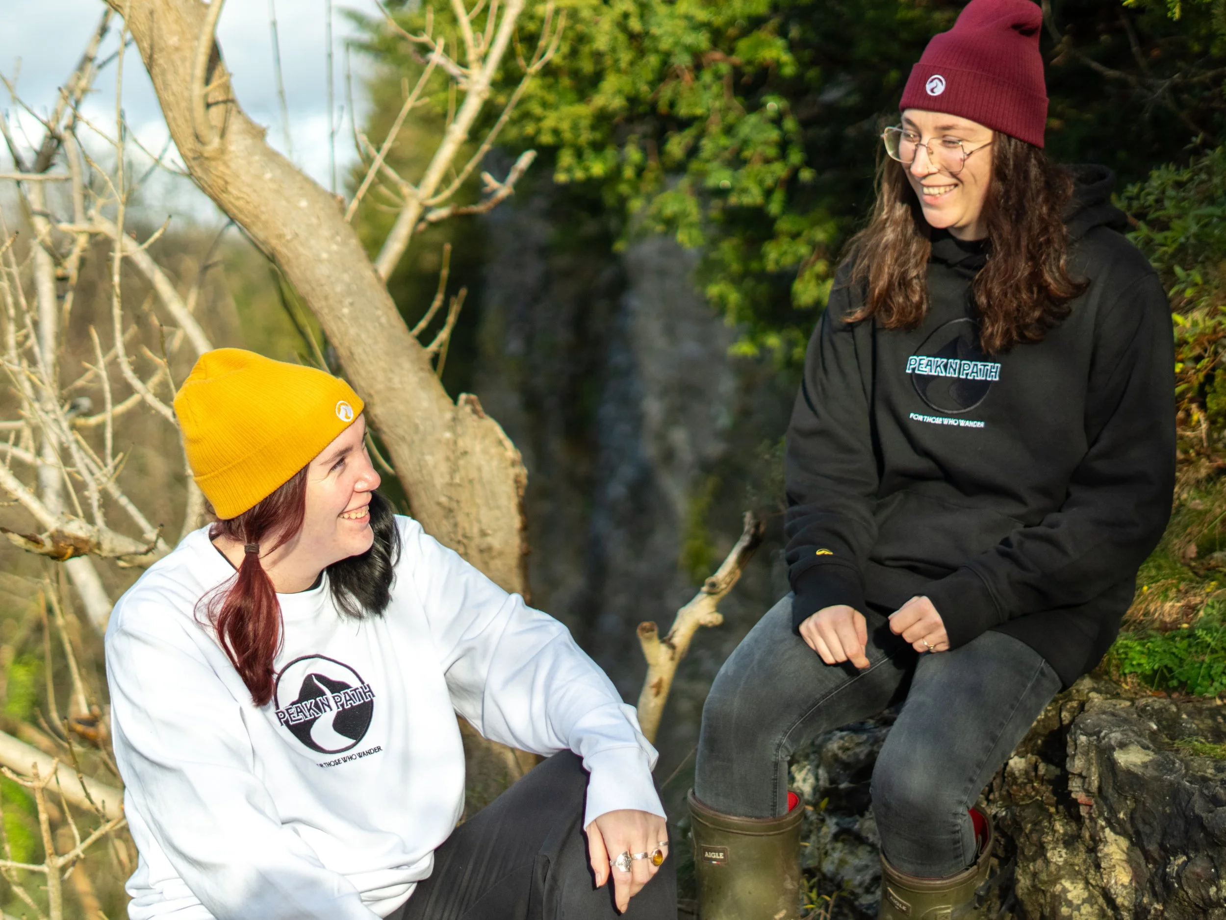Two women sitting outdoors near a tree, smiling and talking. They are wearing beanies and casual outdoor clothing, suggesting they are enjoying a hike or nature outing.