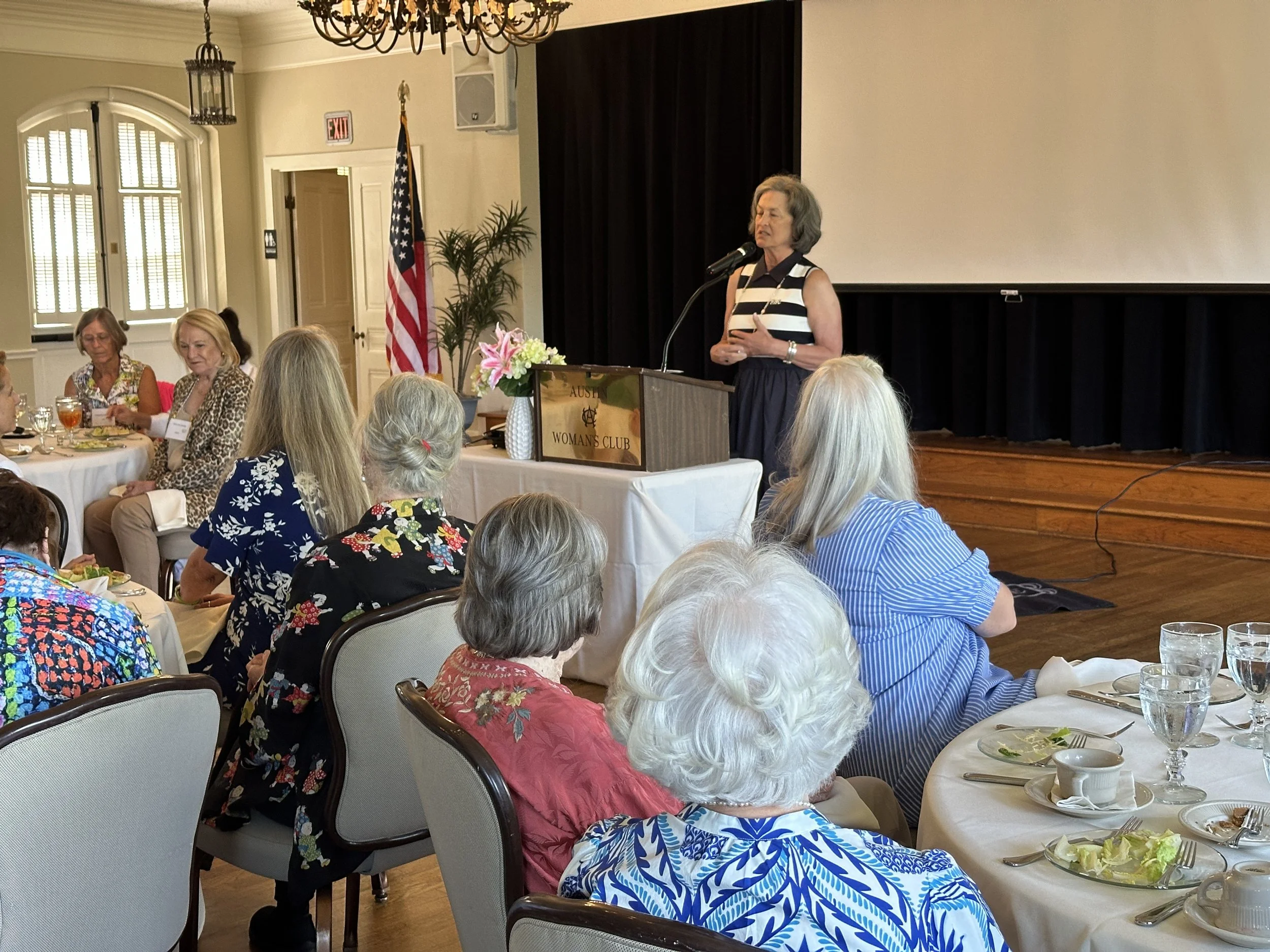 A woman giving a speech at a podium during a luncheon event with elderly women seated at tables decorated with flowers and tableware. A large screen is behind her, and American flags are displayed.