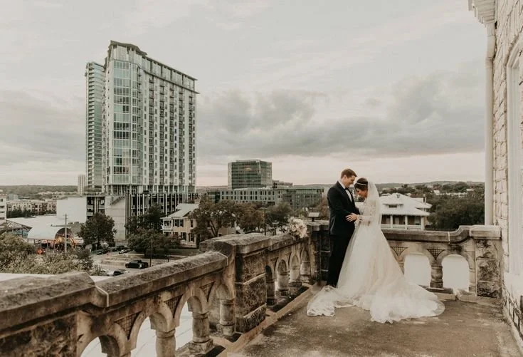 Bride and groom standing together on a balcony overlooking a city skyline with modern buildings, under cloudy skies.
