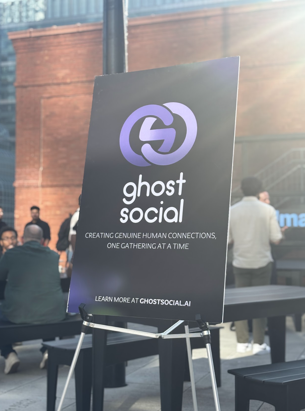 Signboard for Ghost Social, featuring a logo with purple interconnected circles. The sign promotes creating genuine human connections and directs to ghostsocial.ai for more information. In the background, people are gathered in an outdoor urban setting during daylight.