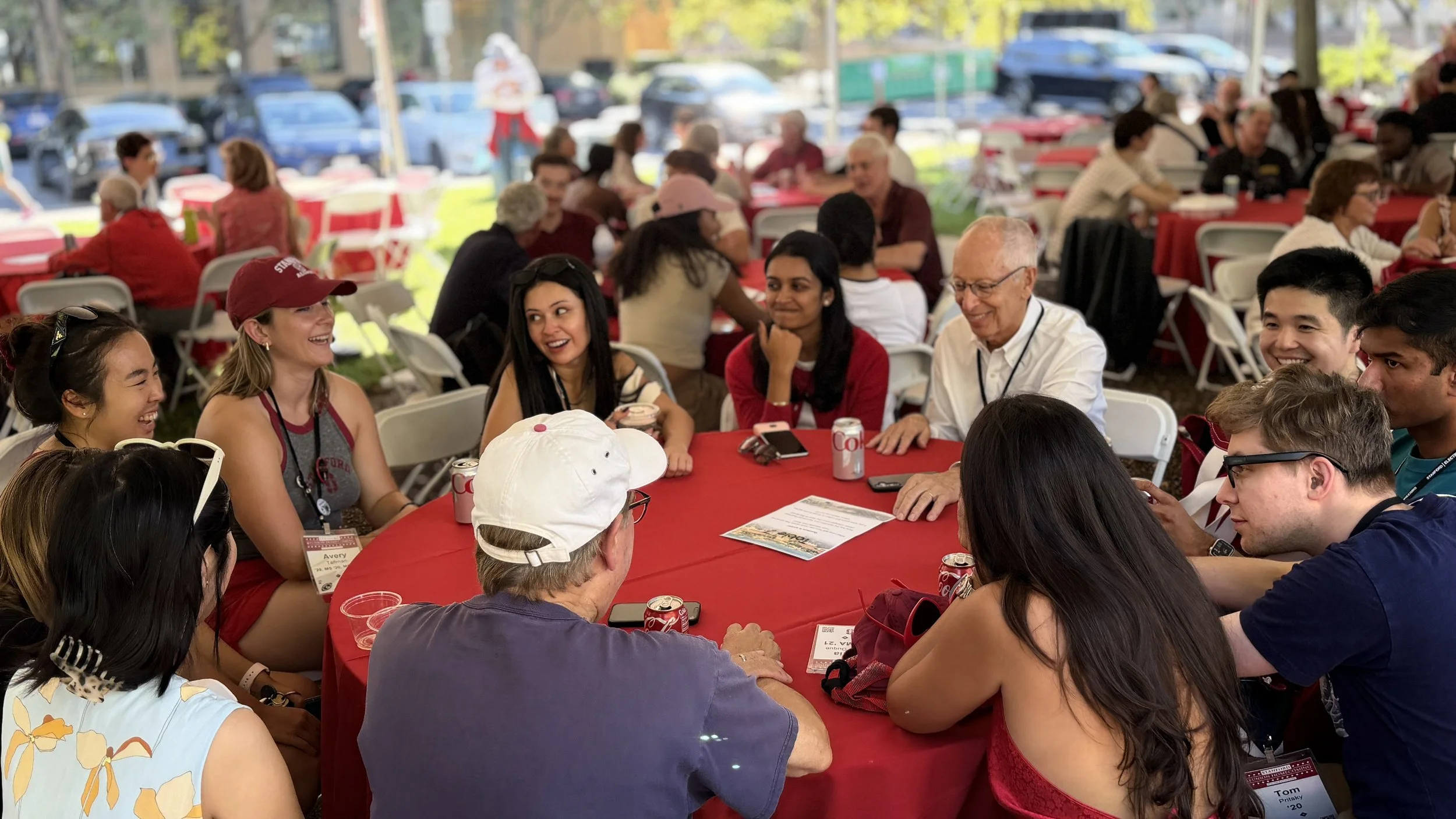 People gathered around a red-covered table outdoors, enjoying a social event or celebration.
