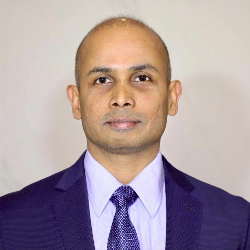 A professional headshot of a man wearing a dark blue suit, light blue shirt, and a patterned blue tie, with a light-colored background.