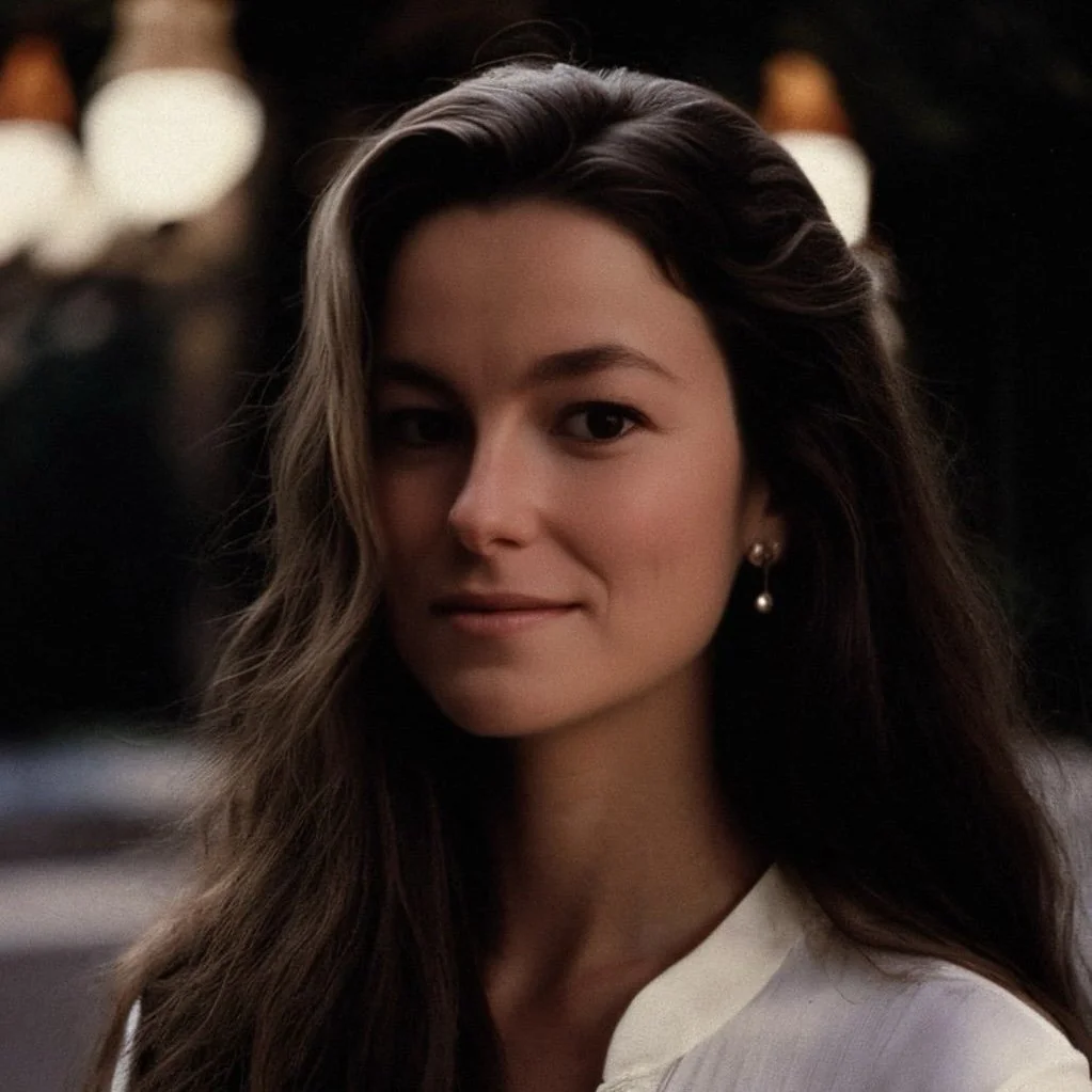 A young woman with long wavy dark hair, wearing a white top and pearl earrings, smiling softly at the camera in a dimly lit setting with blurred lights in the background.