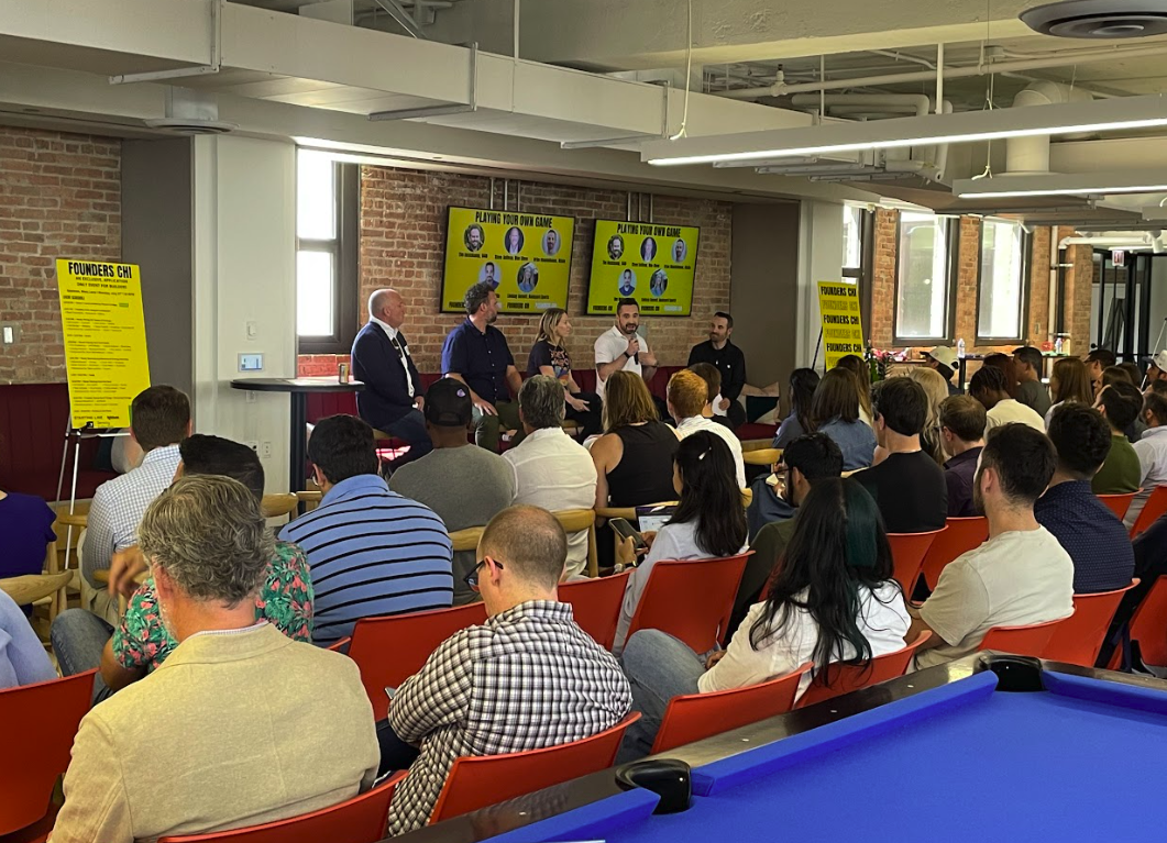 A panel discussion takes place in a room with brick walls and large windows. Five panelists sit on stage facing an audience. The audience is seated in orange chairs, attentively listening. Two yellow posters are visible on either side of the stage, and two screens behind the panel display text and images.