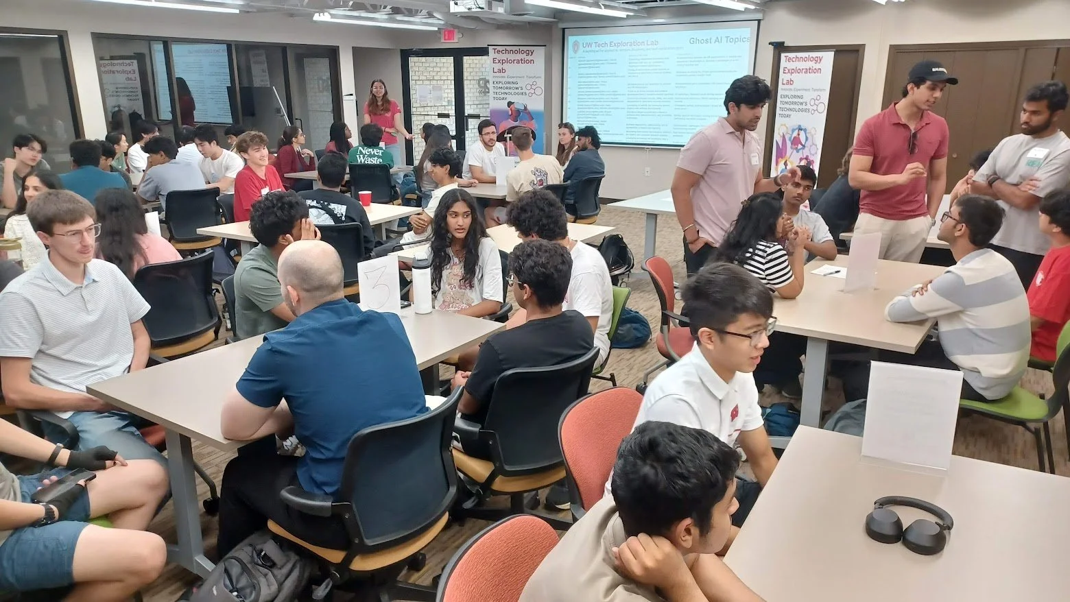 Students and instructors participating in a technology exploration lab, engaging in discussions and presentations with screens displaying information on AI topics.