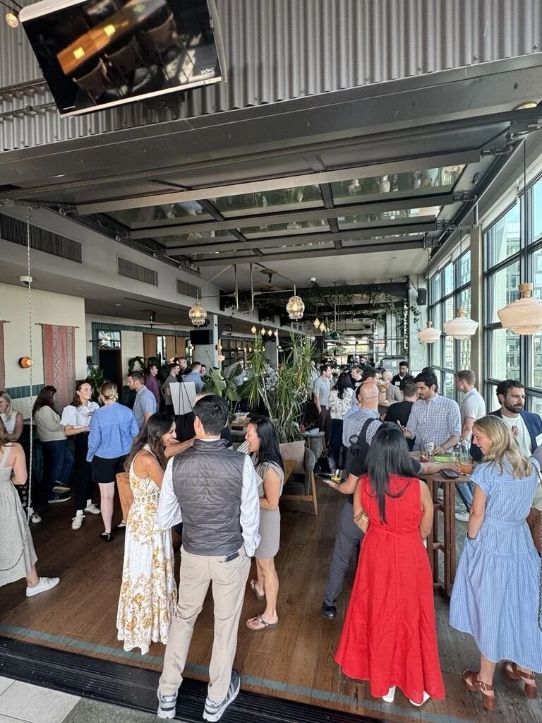 People socializing inside a modern, well-lit restaurant or cafe with large windows, plants, and hanging light fixtures.