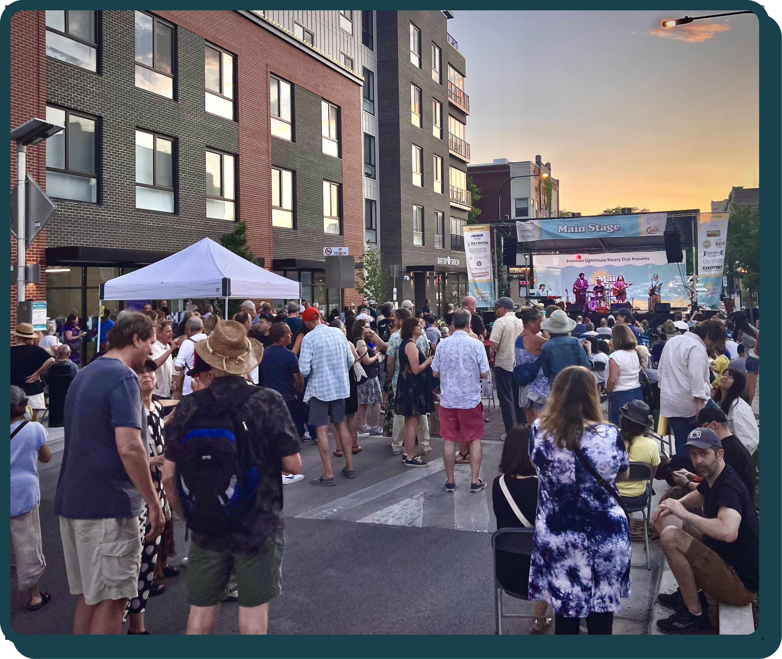Crowd gathered at an outdoor concert stage in an urban area during sunset, with people standing and sitting, watching a live band perform.