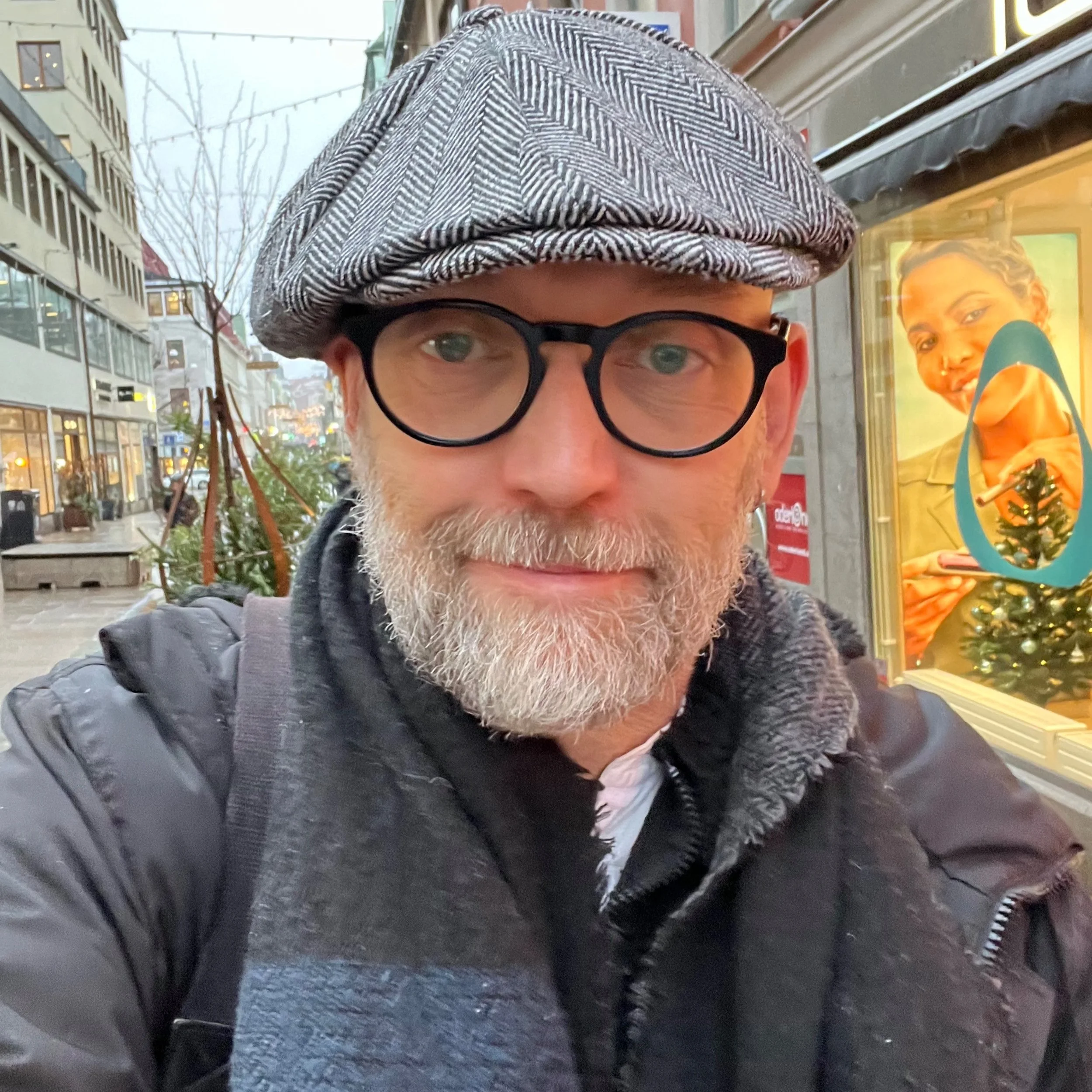 Henrik med glasögon och skägg bär en randig keps och mörk jacka, står på en stadsgata med skyltfönster i bakgrunden.