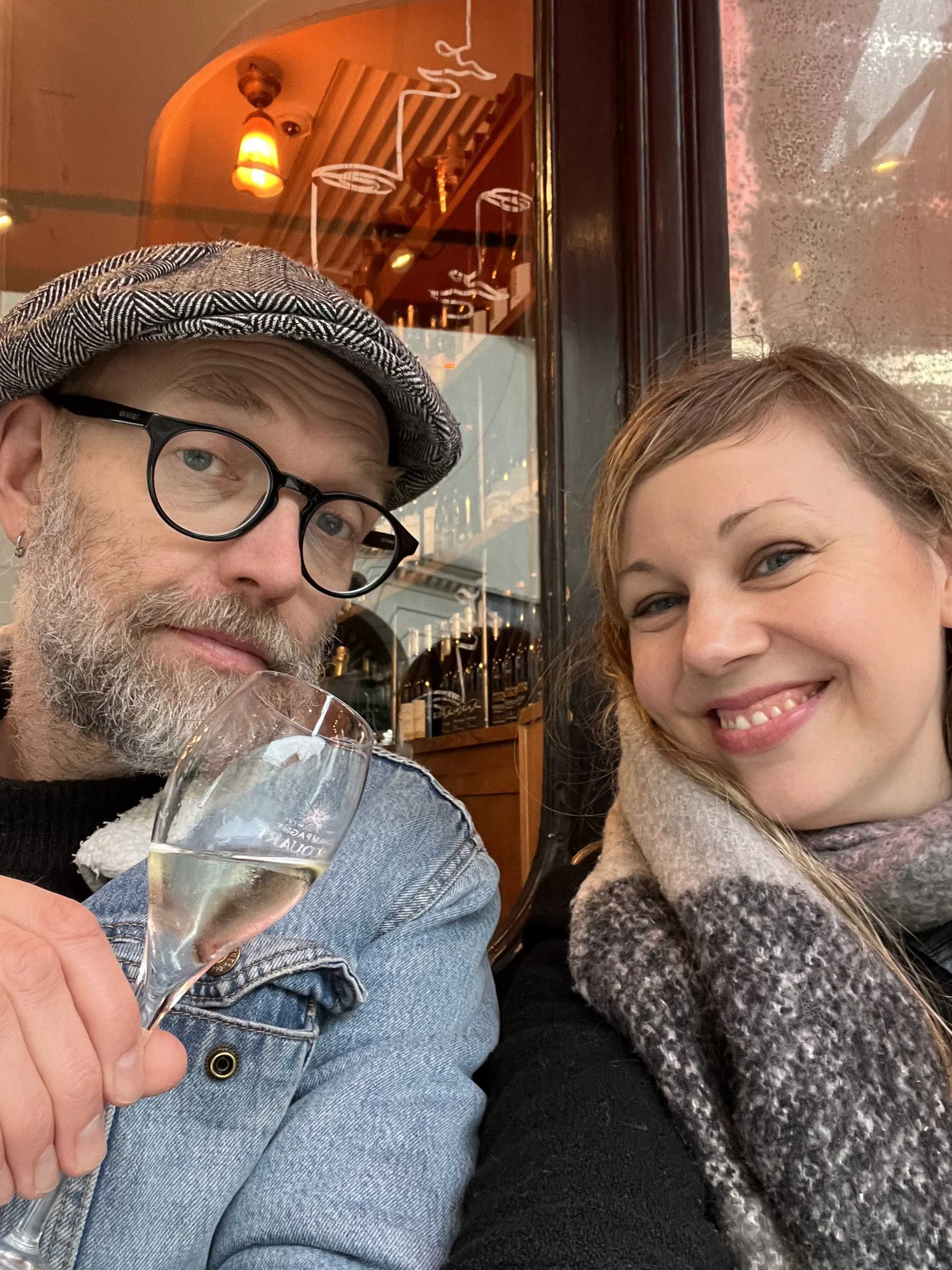 Angelina och Henrik sitter på en restaurang eller ett café. Personen till vänster håller i ett glas med dryck. Båda ser glada ut och ler mot kameran.