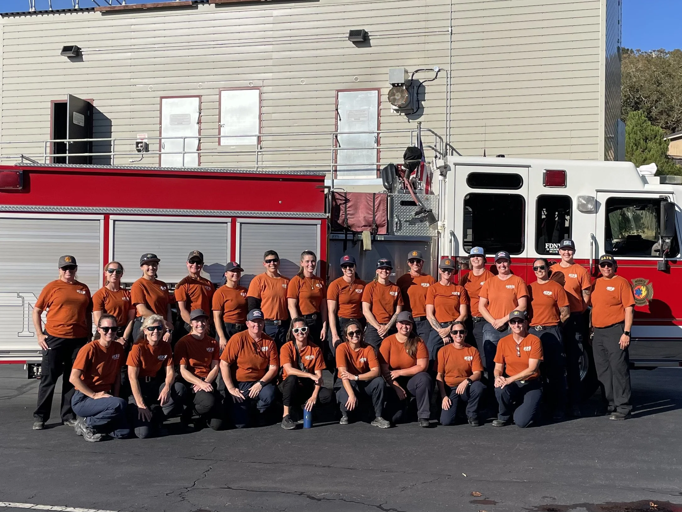 Group of 18 female firefighters in orange shirts posing in front of a fire truck outdoors.