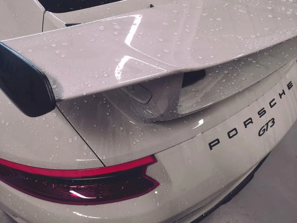 Close-up of the rear of a silver Porsche GT3 sports car with water droplets, showing the Porsche badge and taillight, and a rear spoiler.