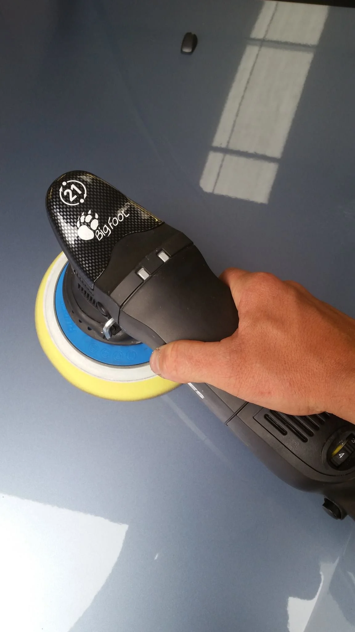 Person holding a bigfoot-brand electric polisher or buffer, polishing a gray car surface.