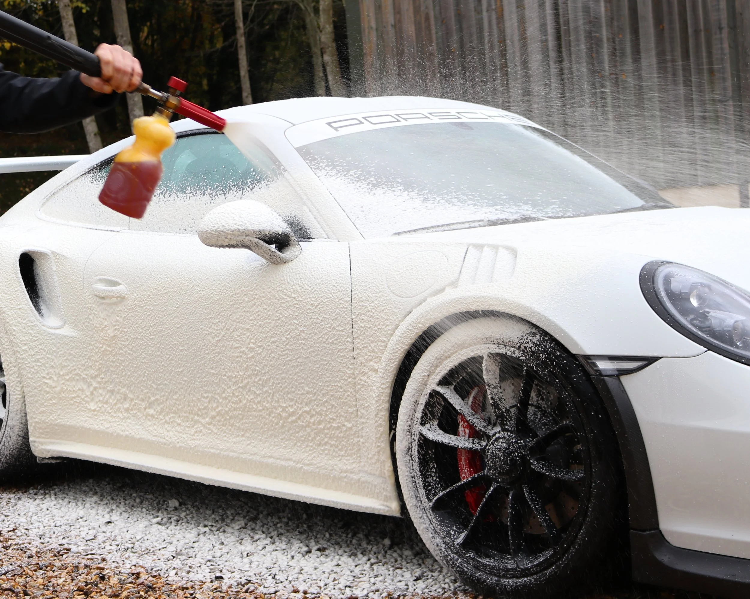 A white Porsche sports car being washed with foam and Pure water, with a person spraying the foam onto the car.