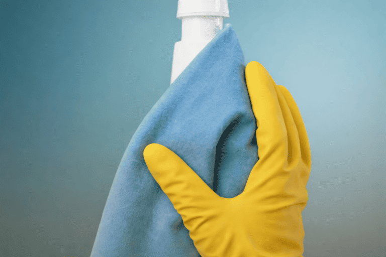 Hand in gelber Gummihandschuh hält einen sauberen, weißen Sprayflaschenkopf mit blauer Reinigungstuch davor gegen einen blauen Hintergrund.