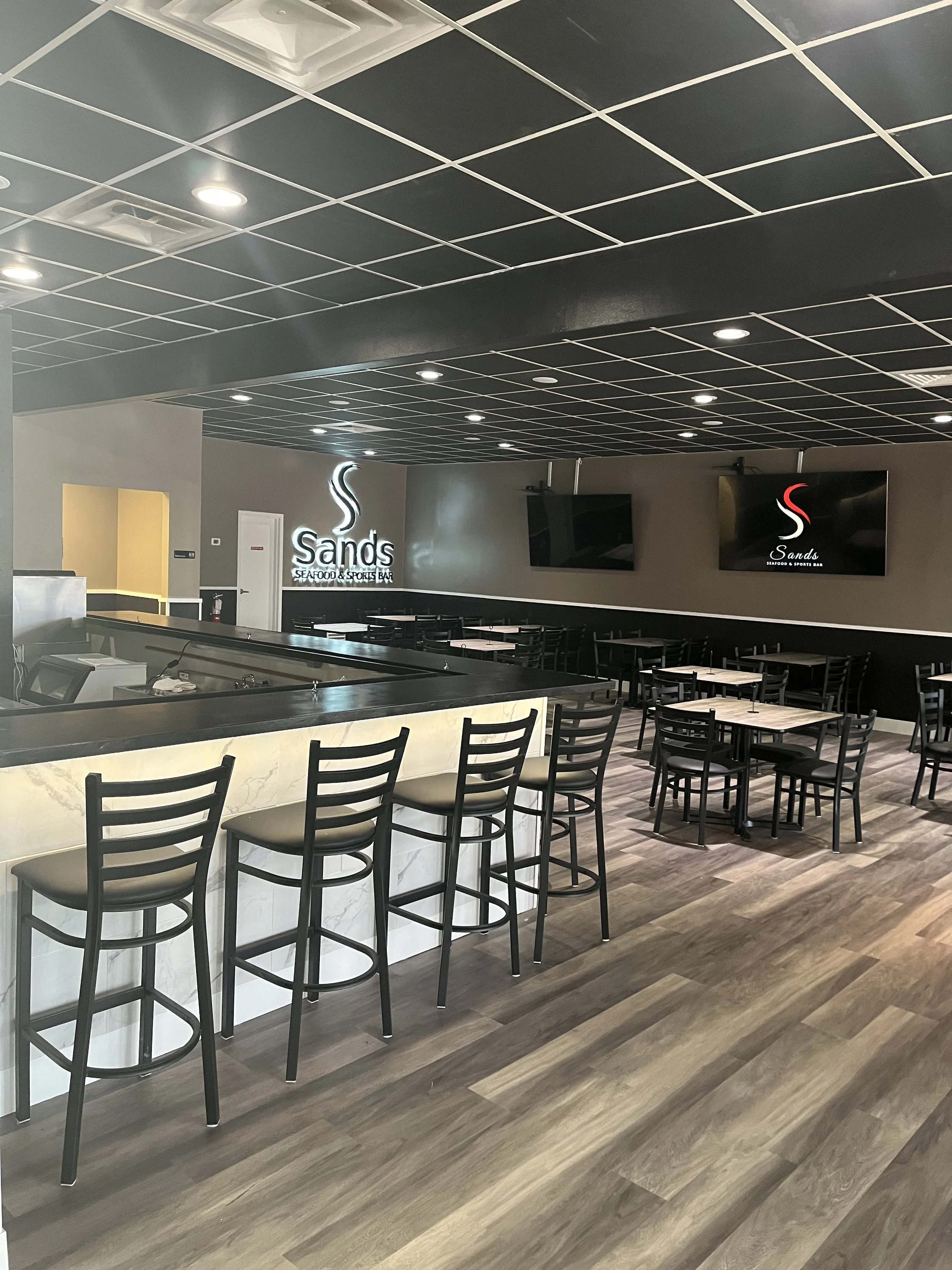 Interior of a seafood and sports bar named Sands with a bar counter and multiple seating tables and chairs, including high chairs at the bar.