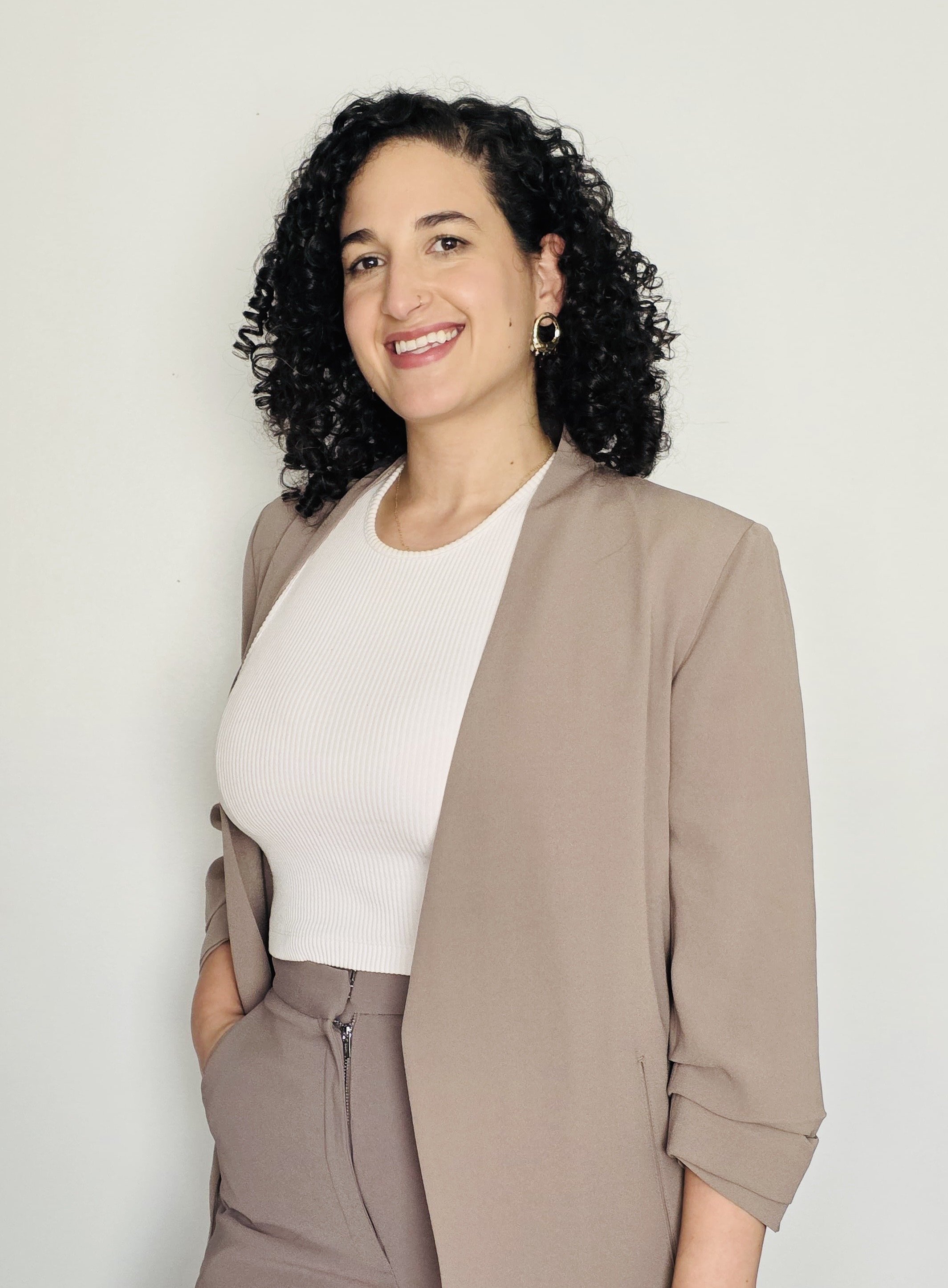 A woman with dark curly hair, wearing a beige blazer over a white ribbed top, standing against a plain white wall, smiling and looking at the camera.