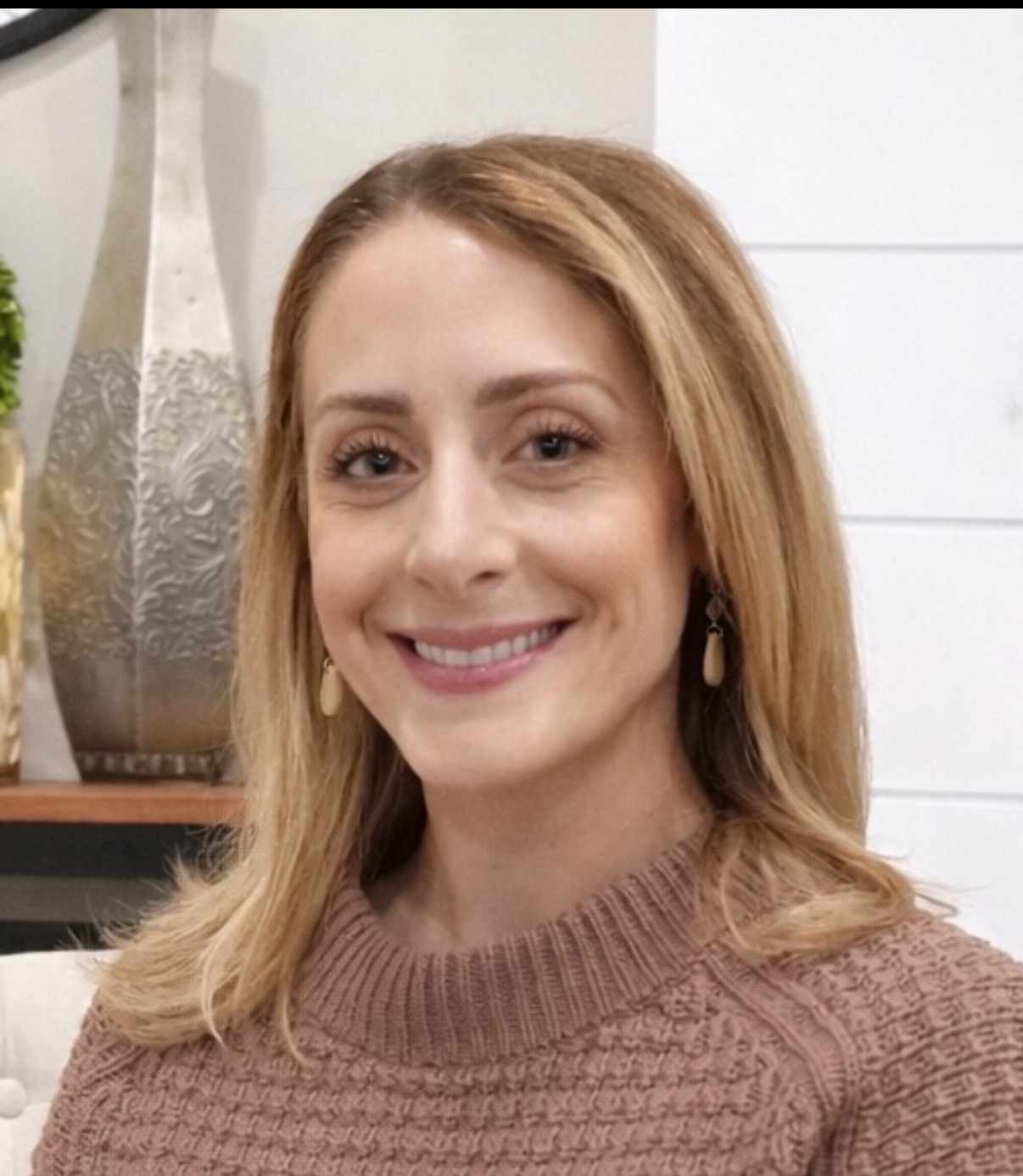 A smiling woman with light red hair, wearing a brown textured sweater and earrings, standing in front of a decorative metallic vase and plants.