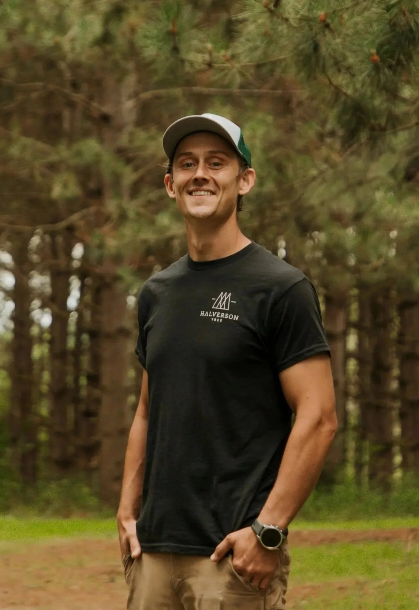A man standing outdoors in a forested area near a body of water, wearing a black t-shirt, a light-colored neck gaiter, a black headband, and a watch, smiling at the camera.
