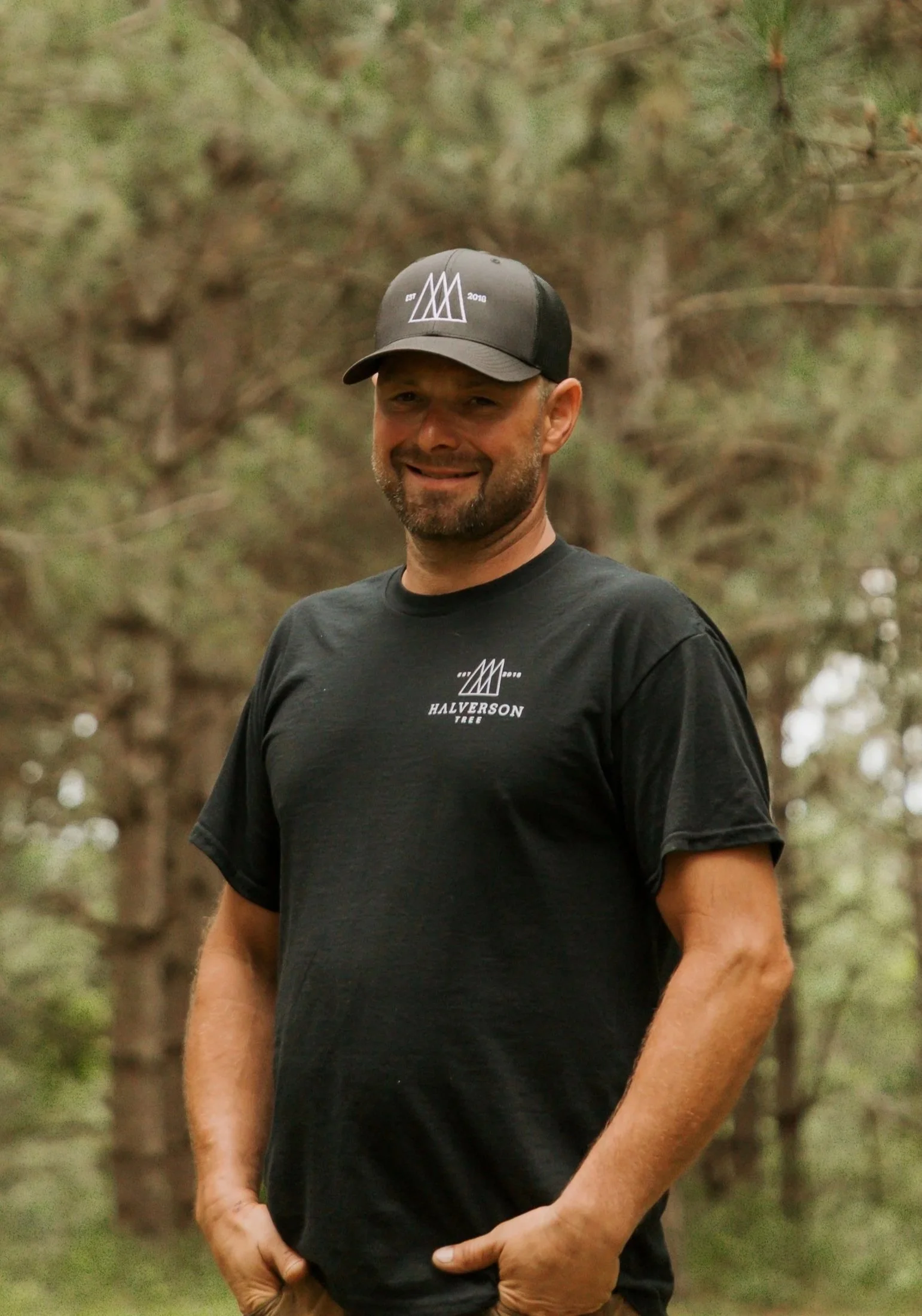 A man with a beard smiling outdoors, wearing a black shirt with a logo and text reading 'Halverson Tree' and a baseball cap, standing in front of a river and autumn trees.
