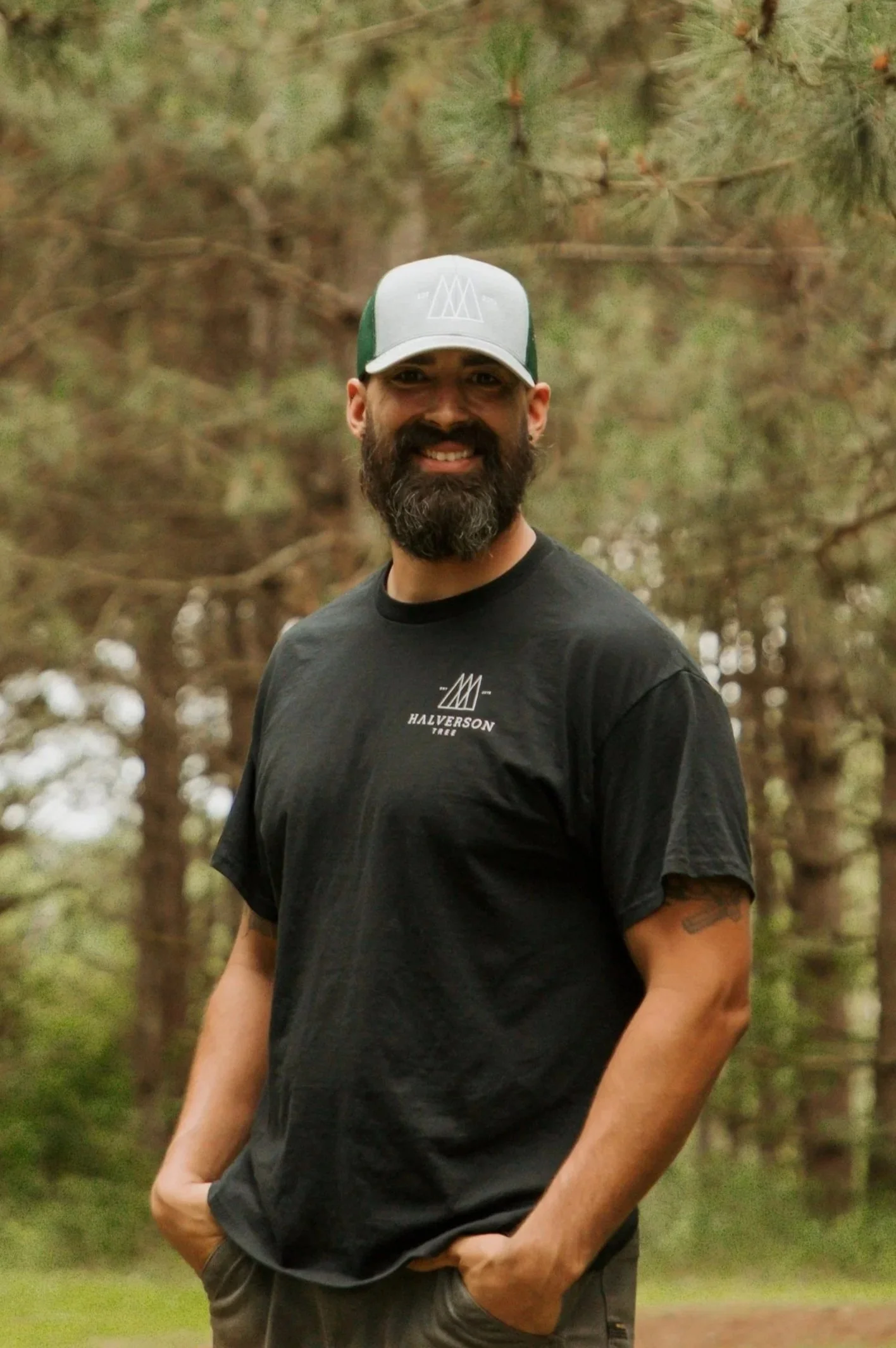A bearded man wearing a gray cap and black long-sleeve shirt standing outdoors near a lake or river with trees and hills in the background during autumn.