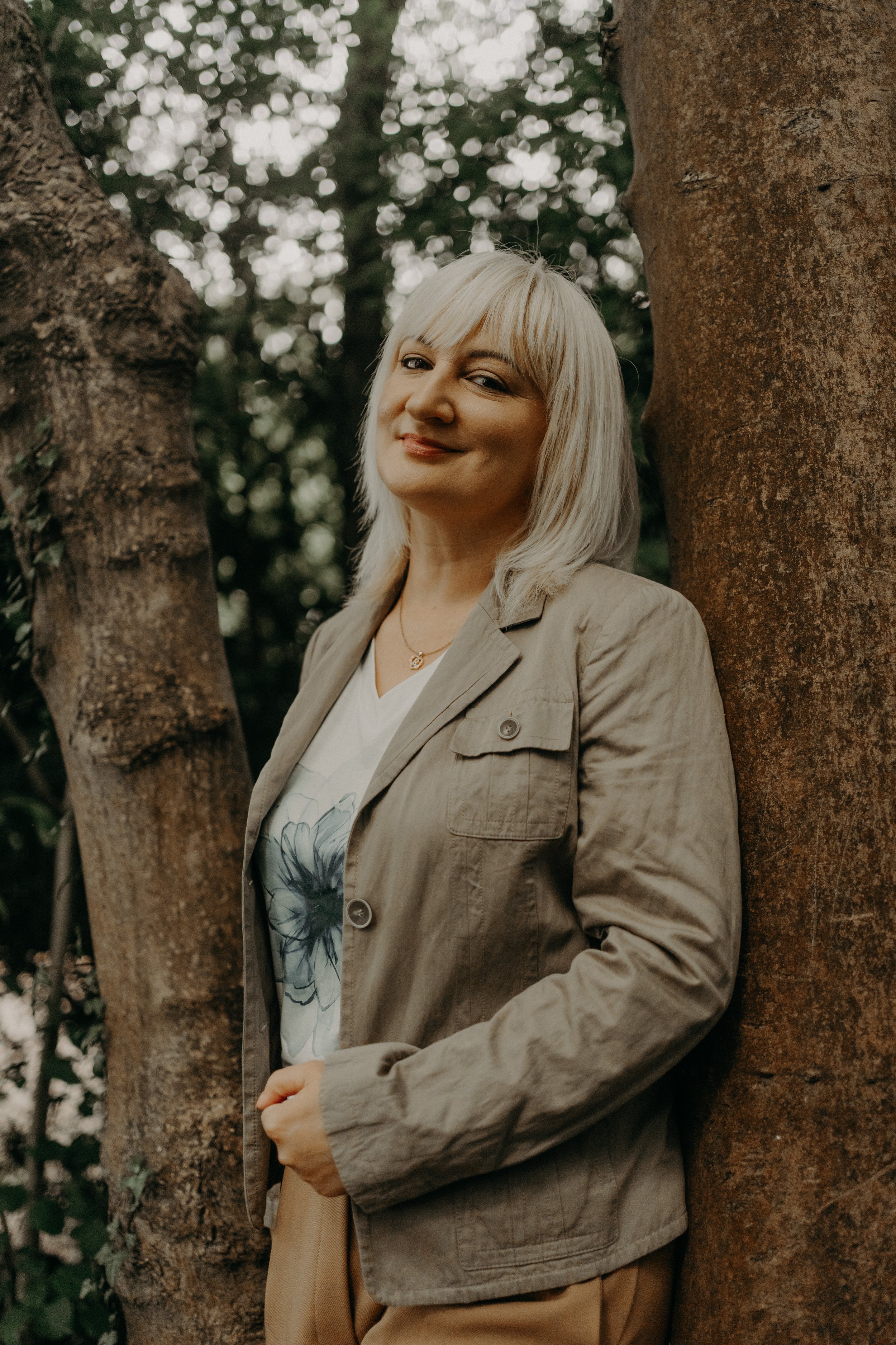 A woman with blonde hair smiling, leaning against a tree in an outdoor setting surrounded by greenery.