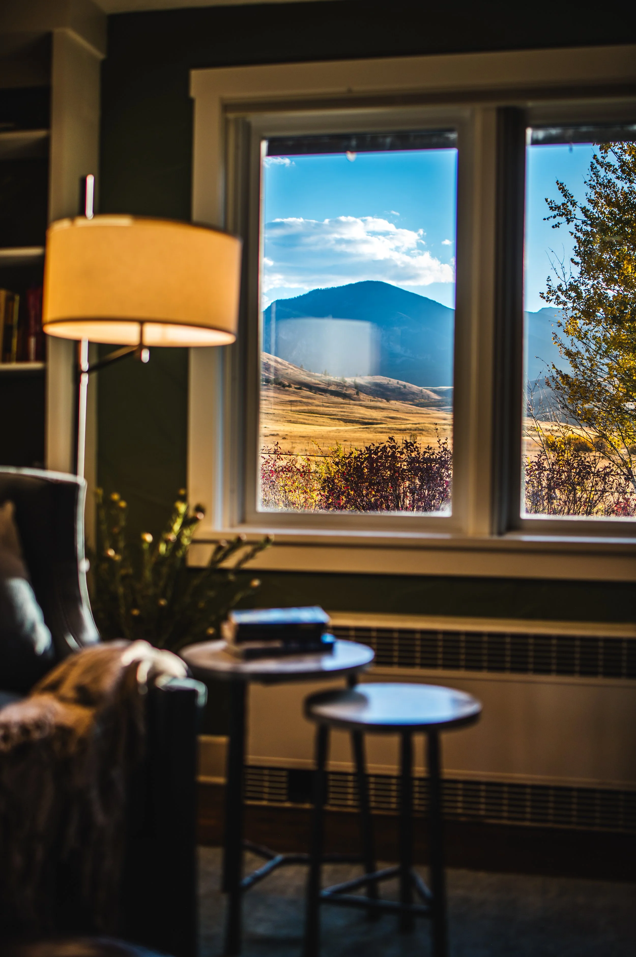Bedroom with large windows overlooking Montana trees, wooden dresser with lamps, blue upholstered armchair, and layered bedding in a Western Roots ranch home, showcasing modern rustic interiors and western home decor in a generational log house.