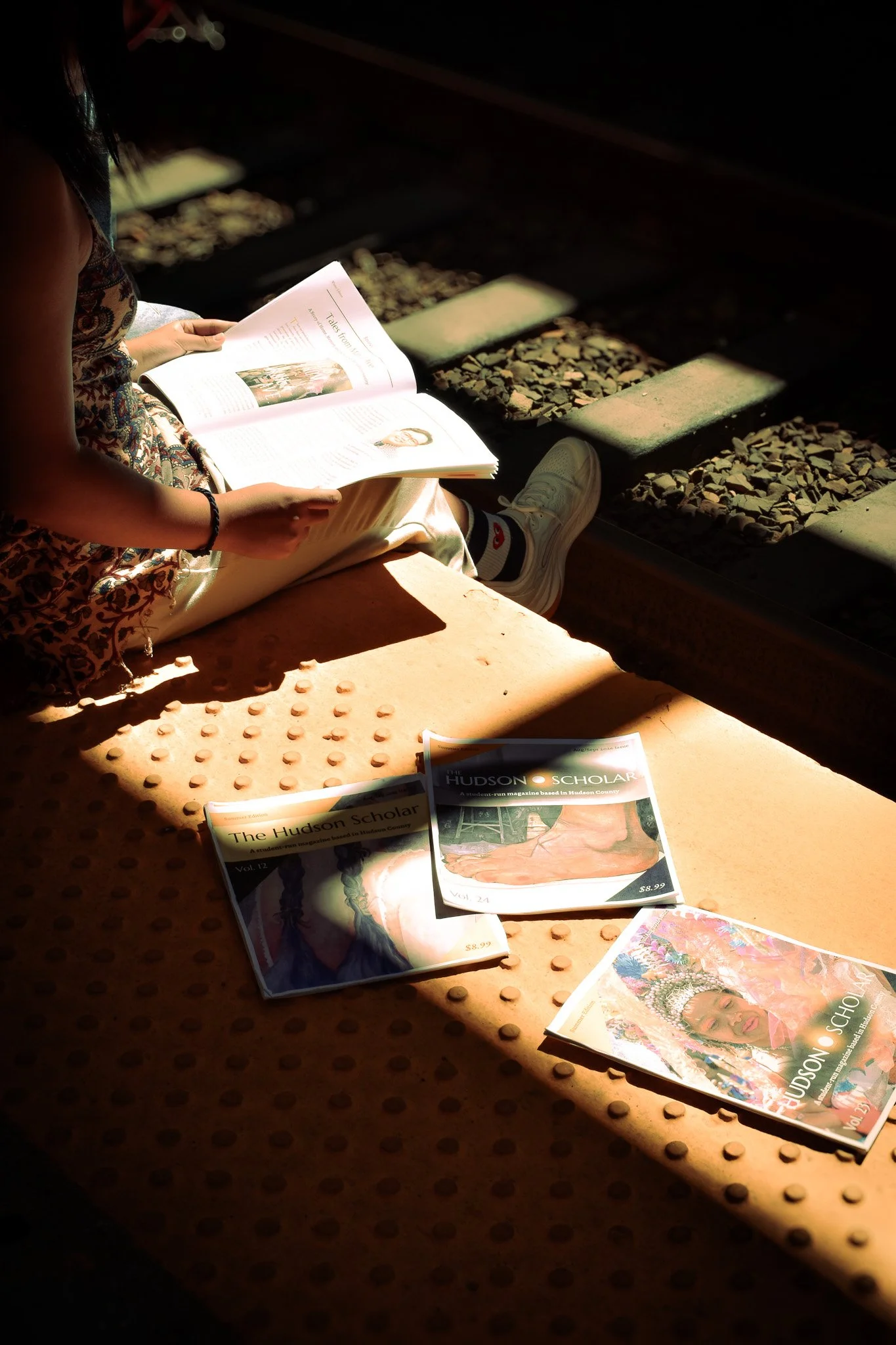 A person sitting on a train platform reading a magazine. Several magazines titled "The Hudson Scholar" are spread out on the ground nearby, under a patch of sunlight, alongside gravel and concrete tiles.