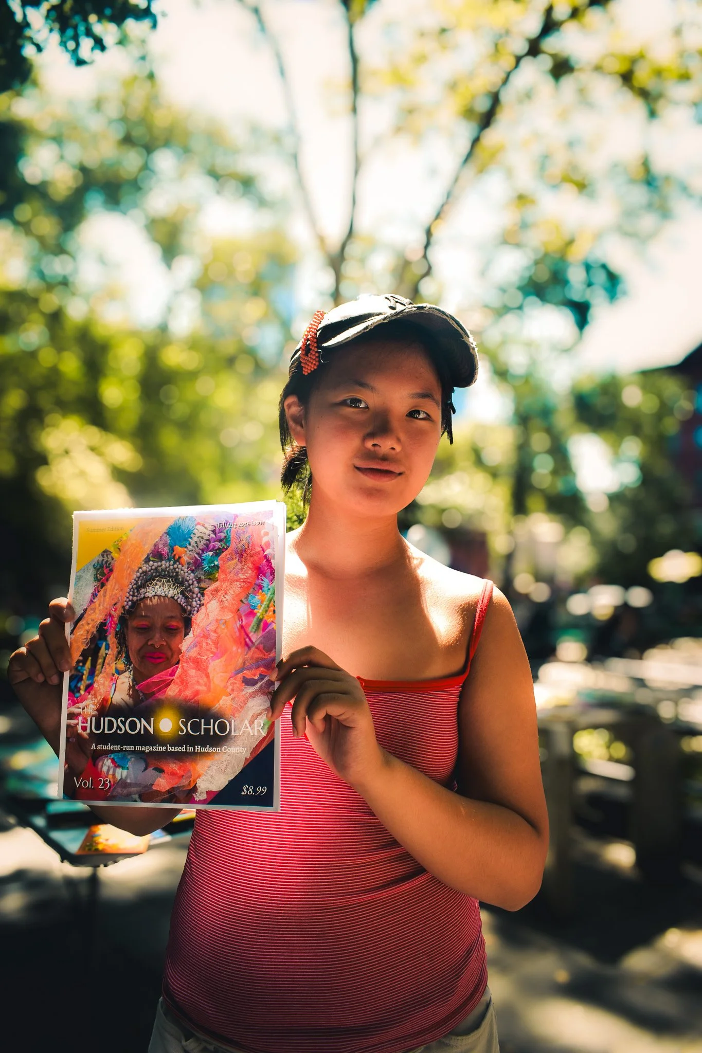 A young woman in a red striped tank top and tan shorts stands outdoors holding a magazine titled "The Hudson Scholar" with a colorful cover image. She is wearing a baseball cap and has short black hair. The background is blurred with trees and sunlight filtering through.
