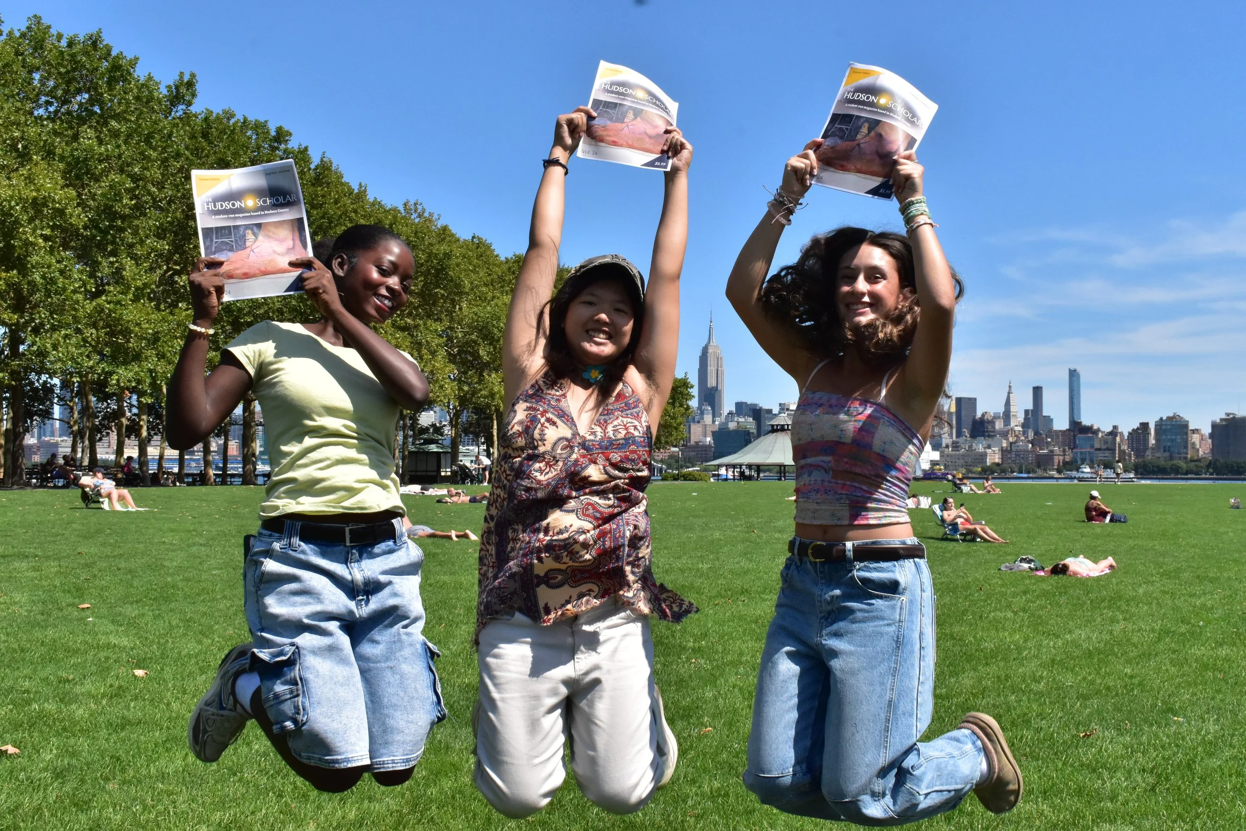 Three young women kneeling on a grassy park with a city skyline in the background, holding newspapers and smiling.