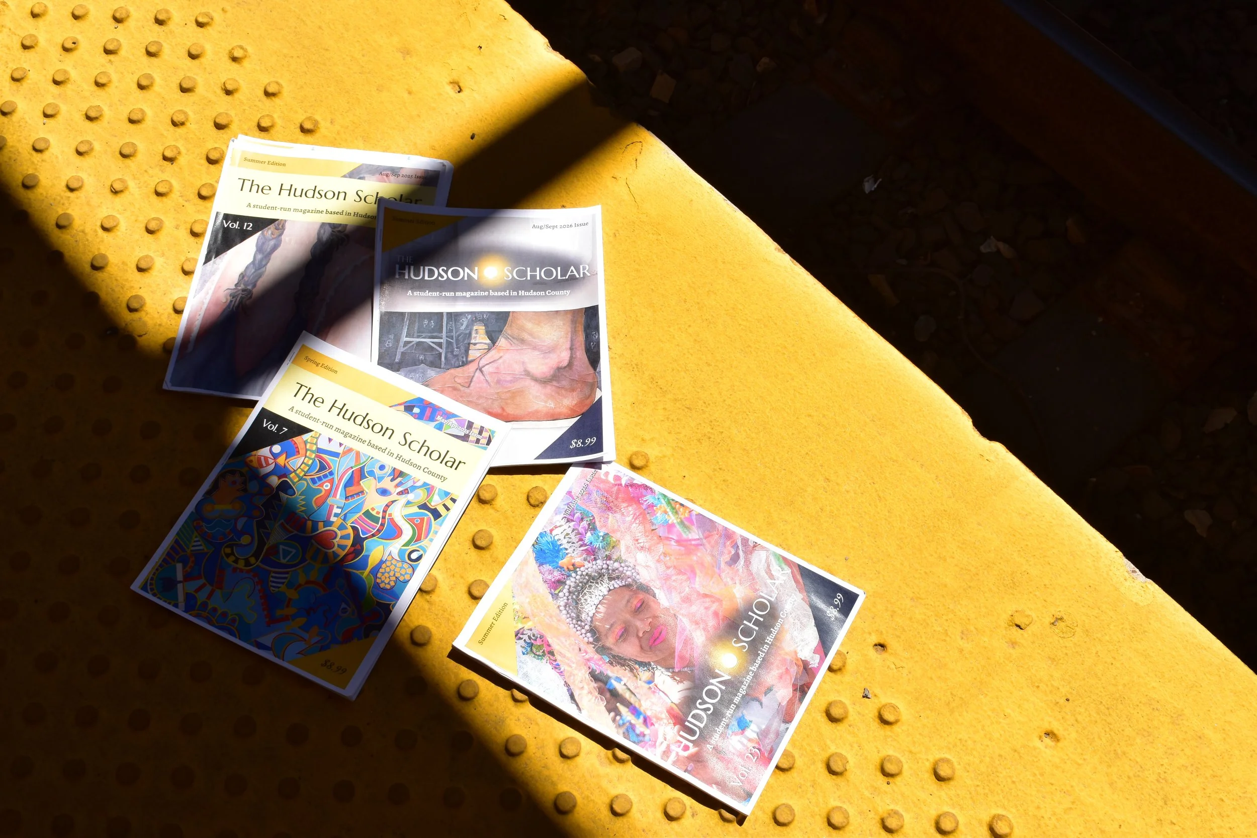 Several issues of 'The Hudson Scholar' student-run magazine placed on a textured yellow surface, with some issues overlapping and a shadow cast over part of the magazines.