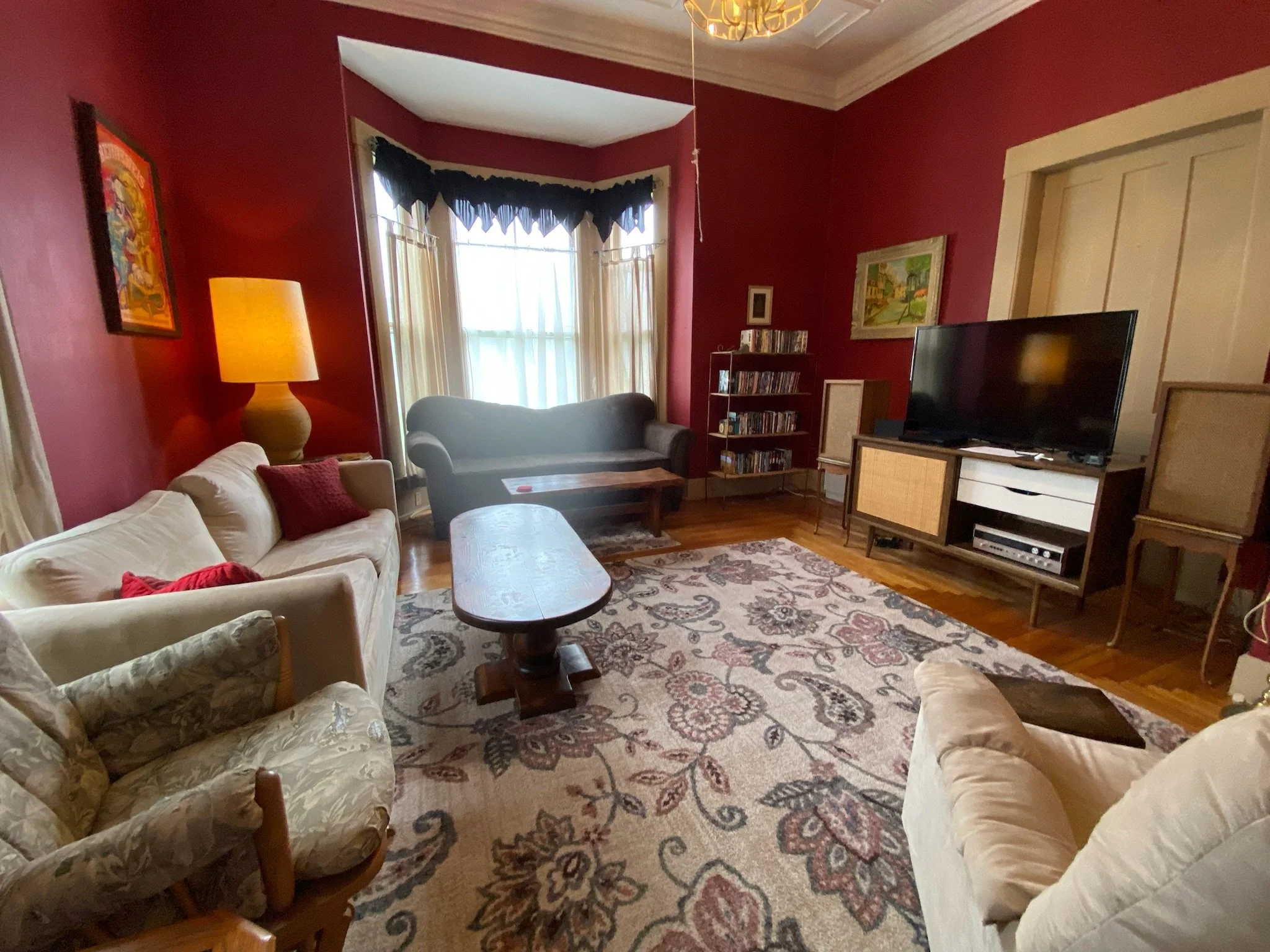 Living room with red walls, a white sofa with red pillows, floral armchairs, a wooden coffee table, a large patterned area rug, a TV on a stand, a bookshelf with DVDs, a window with yellow curtains, a dark valance, a table lamp, and framed artwork on the walls.