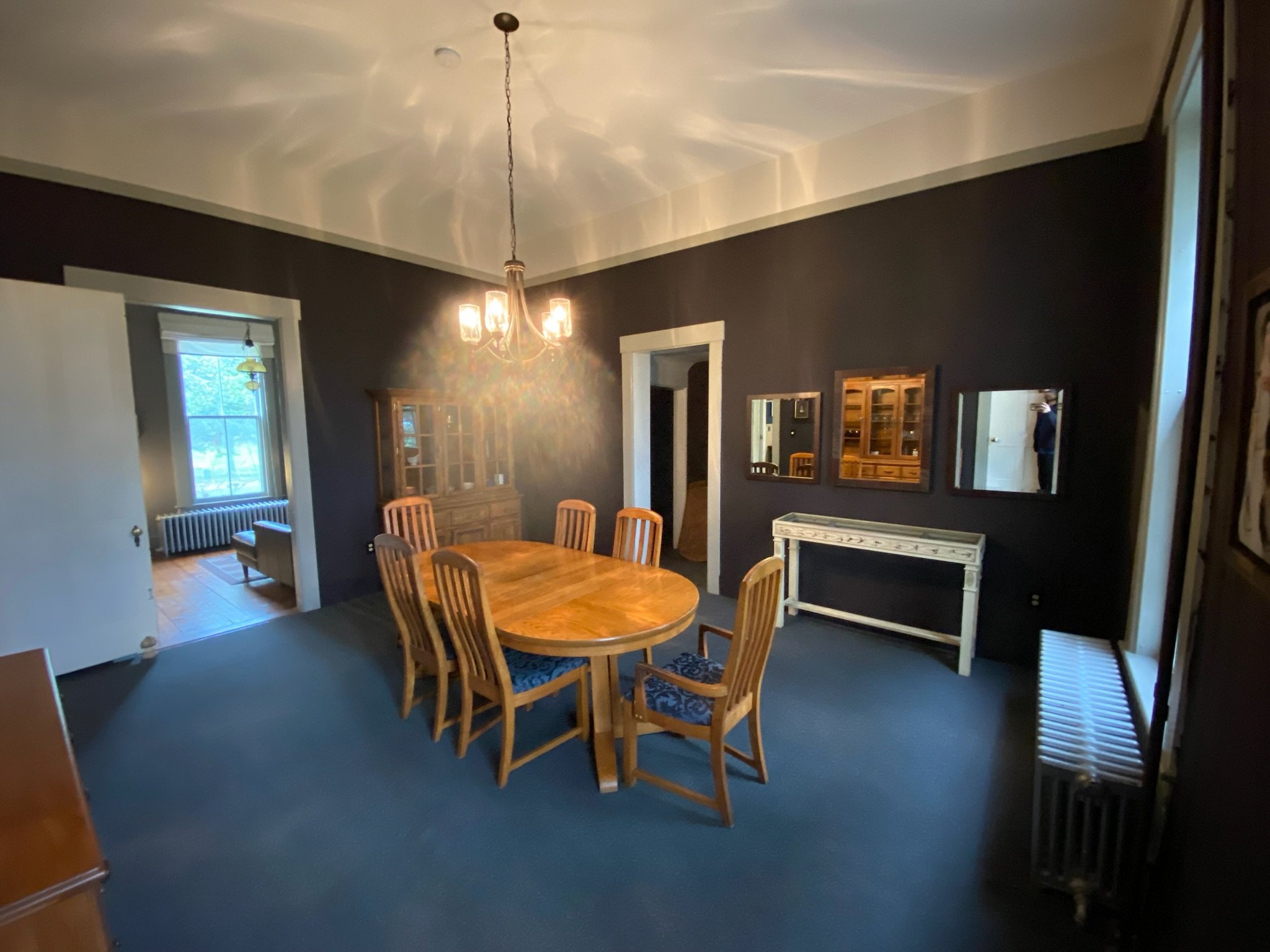 A dining room with a wooden table surrounded by eight wooden chairs with blue cushioned seats. There is a dark wall with three mirrors and a small white console table. A ceiling light fixture with four bulbs and glass shades is hanging from the ceiling. The room is lit by natural light from a window on the left and a radiator below the window.