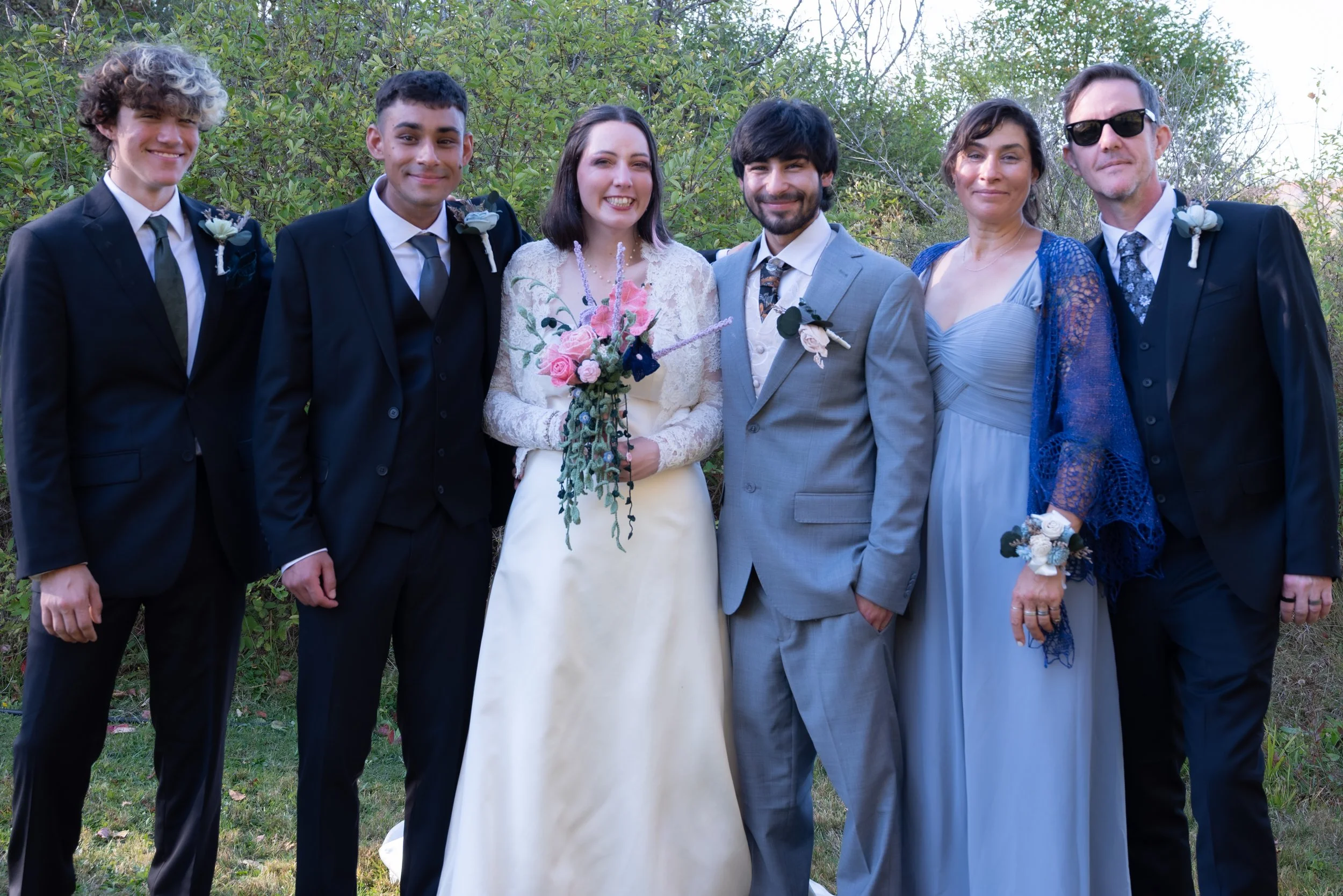 A group of seven people dressed in formal wedding attire standing outdoors with trees and greenery in the background, smiling for a photo.