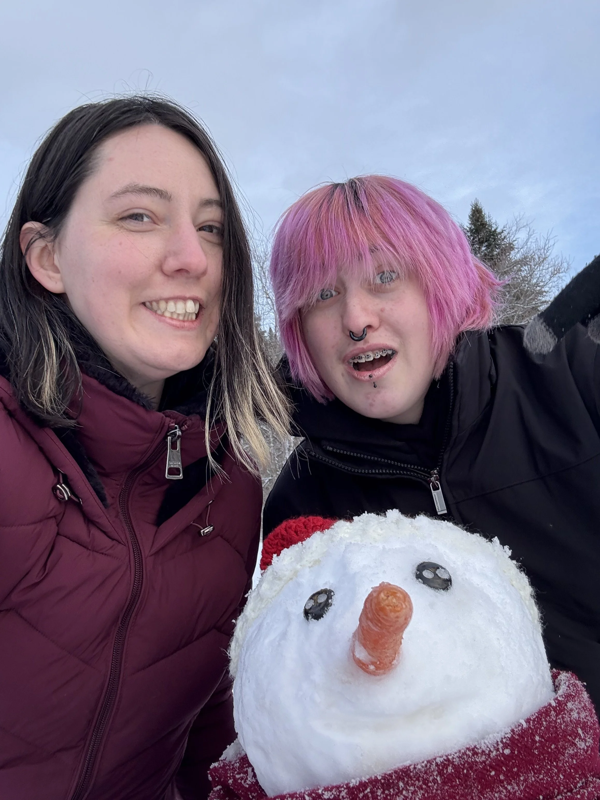 Two people smiling outdoors in winter with a snowman that has a carrot nose and black buttons for eyes.