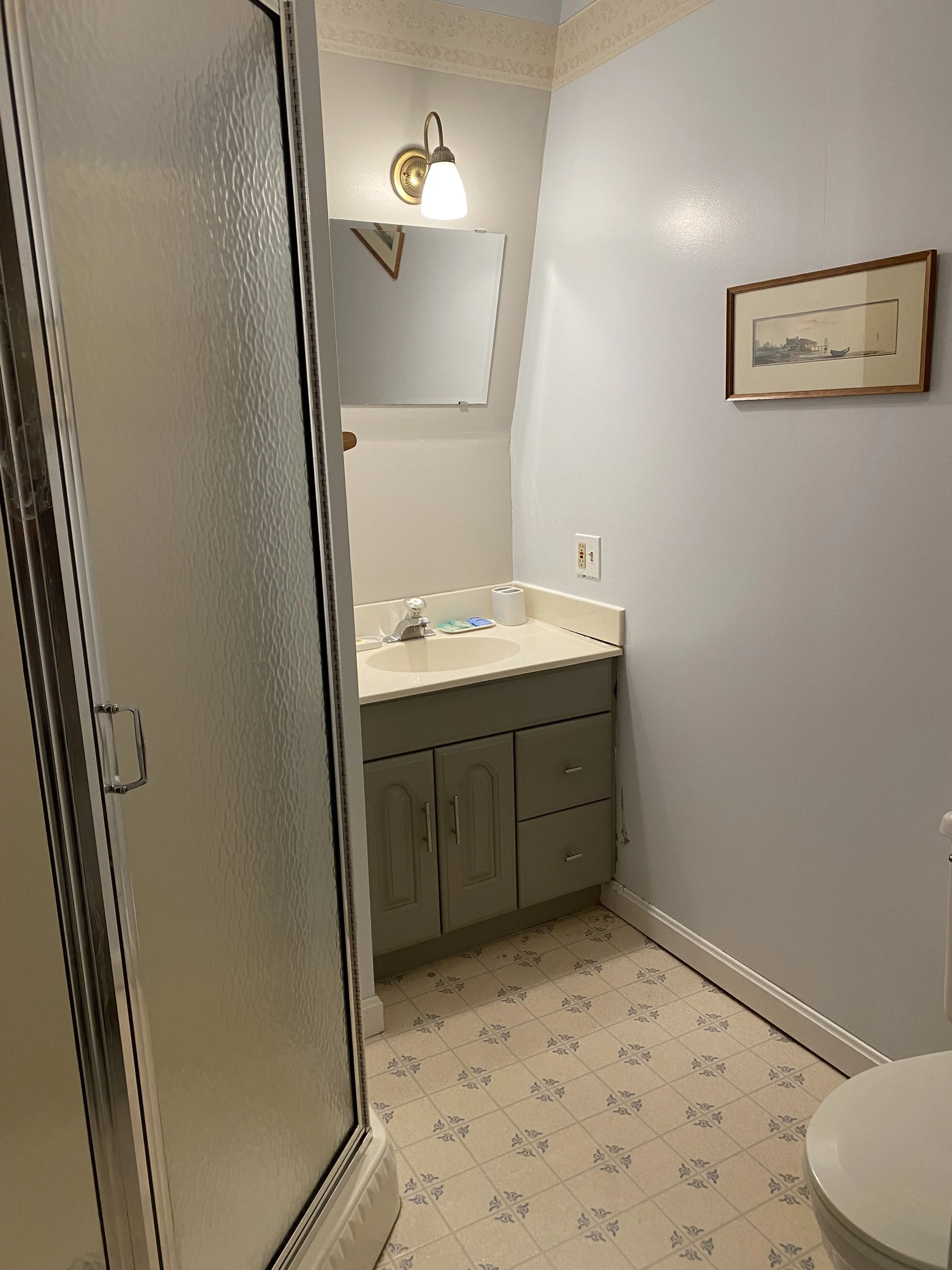 A small bathroom with a toilet, a sink with a cabinet, a mirror, a window with blinds, and a shower stall. There are toiletries on the toilet tank and on the sink.